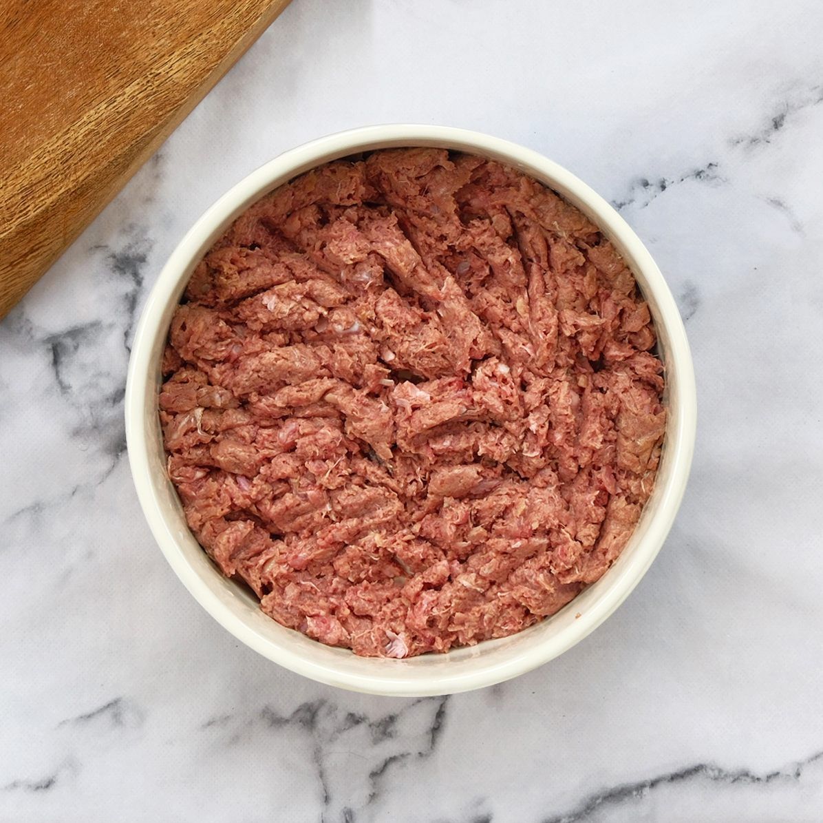A white bowl filled with pinkish-brown, ground meat on a marble surface, a wooden cutting board in the background.
