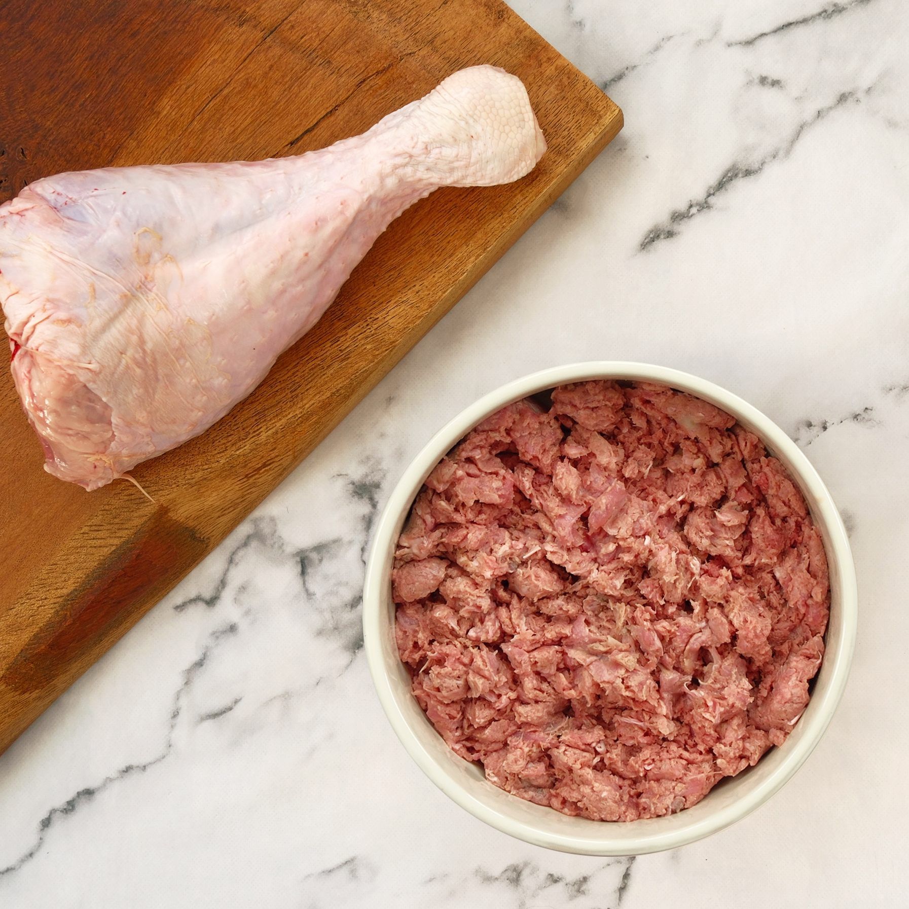 Raw turkey drumstick on a cutting board, bowl of ground meat on marble surface.