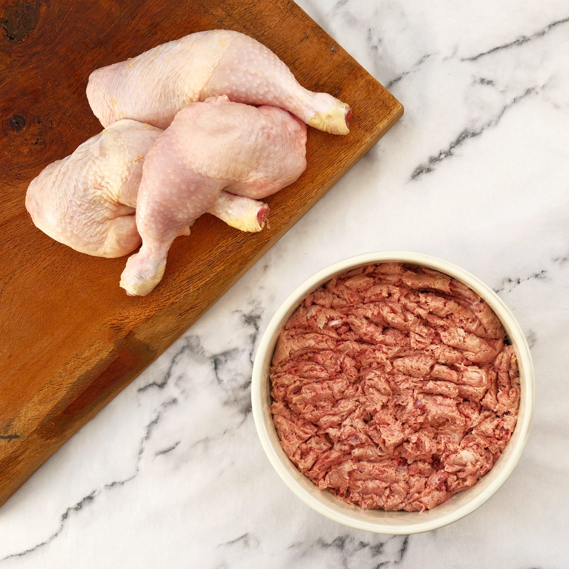 Raw chicken leg quarters on a wooden cutting board next to a bowl of ground raw meat on a white marble surface.