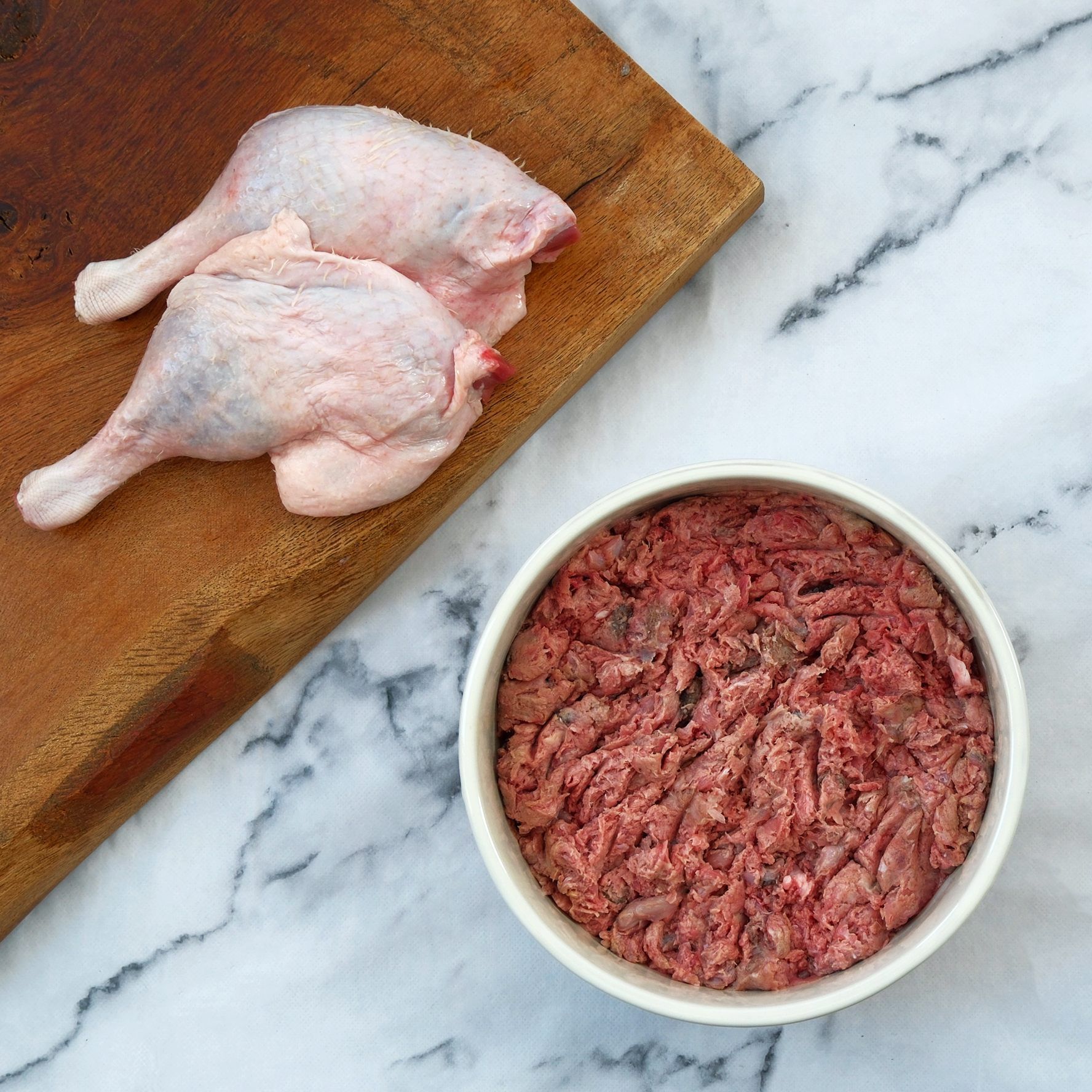 Two raw duck legs on a wooden cutting board next to a bowl of ground raw meat on a marble surface.