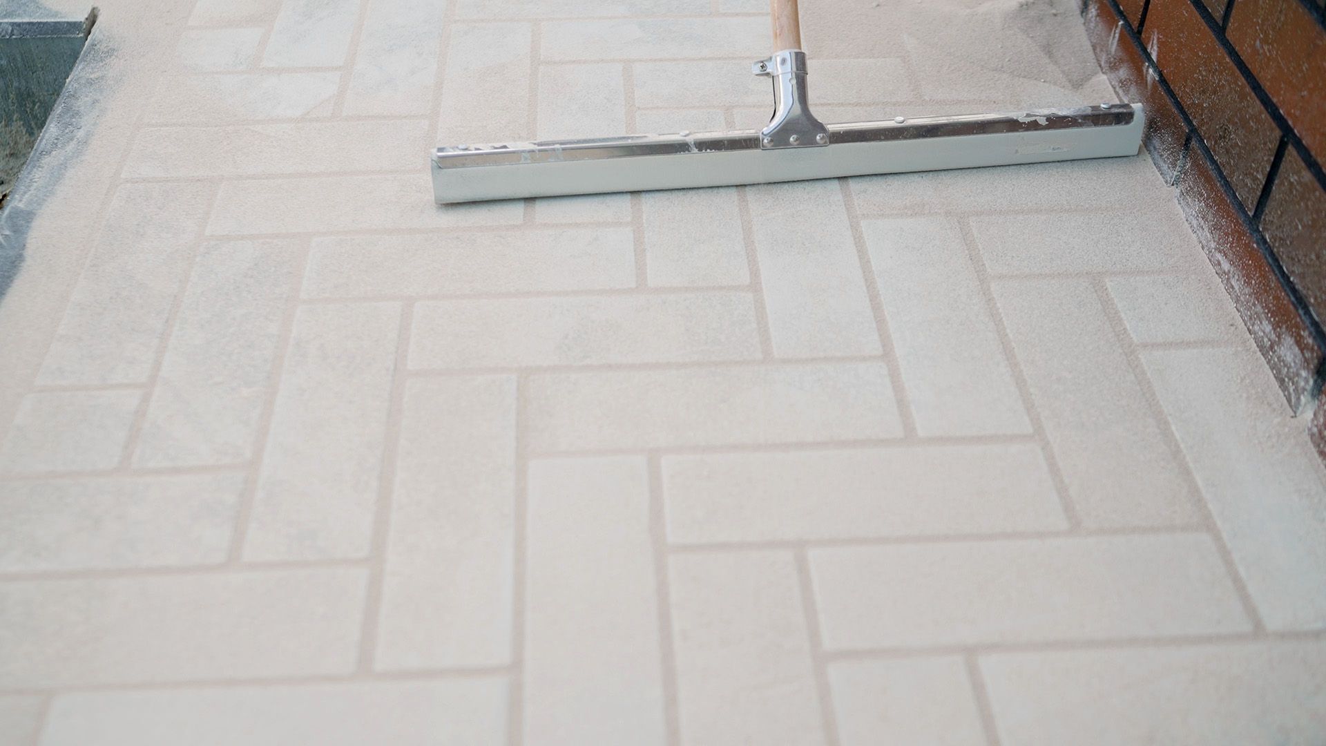 A squeegee spreading sand across freshly laid patio paver tiles.