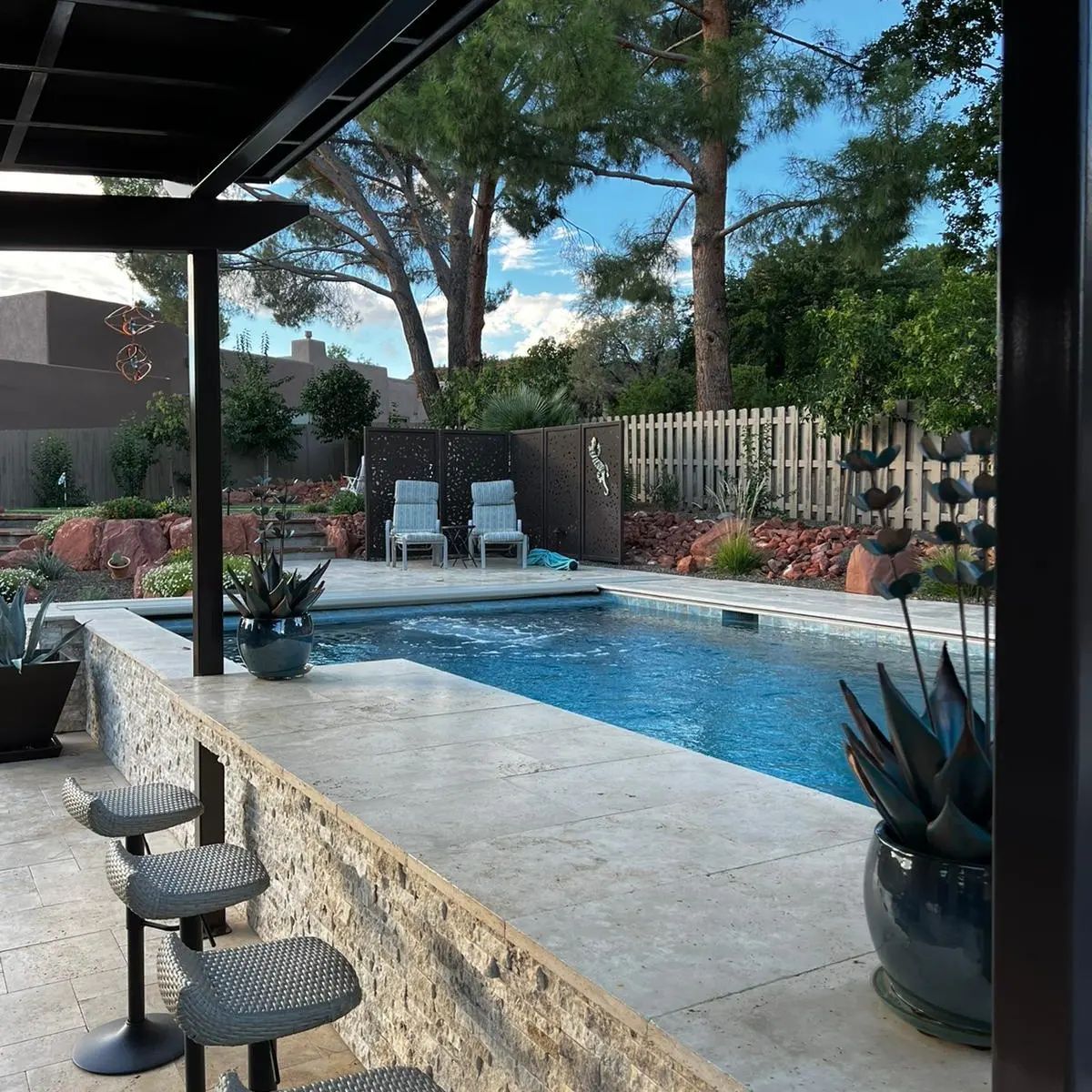 A pool with a stone bar, seating, and lounge chairs in a backyard with trees and a fence.