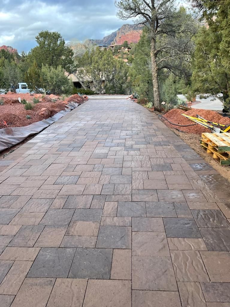 Brick paved driveway leading towards trees and a house, surrounded by landscaping.