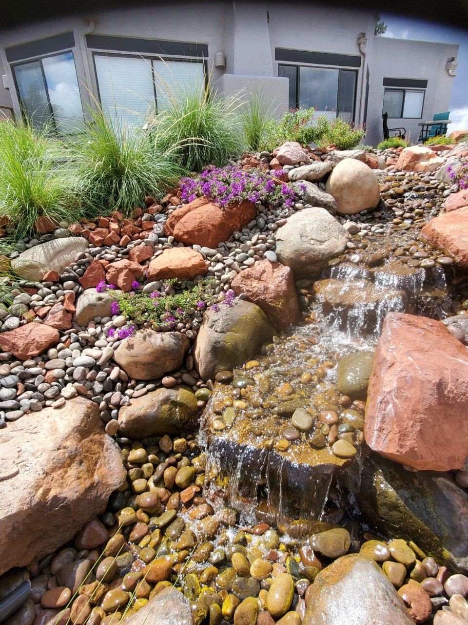 A waterfall surrounded by rocks and flowers in front of a house