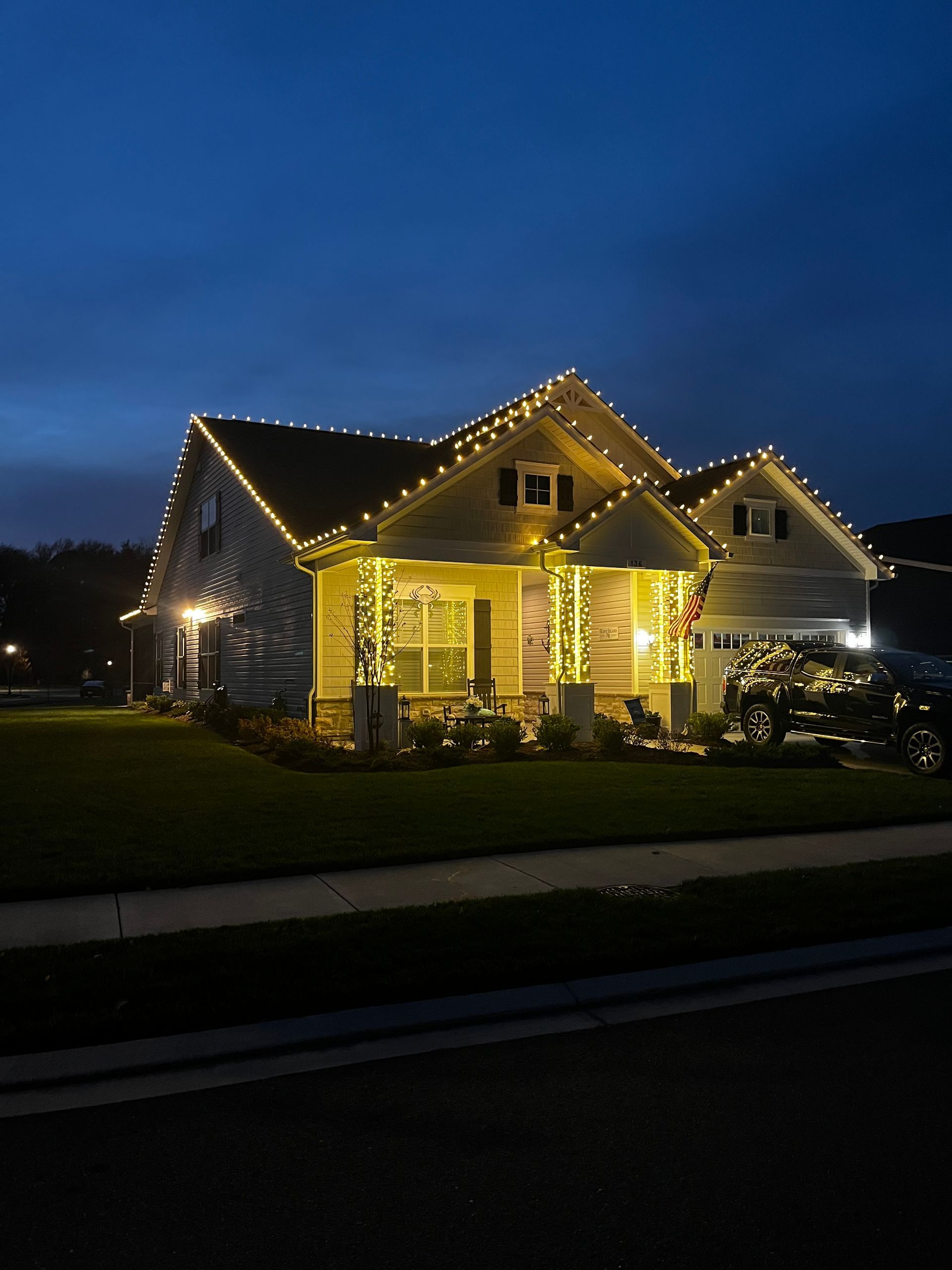 A residential house at twilight decorated with warm yellow string lights along the roofline and porch columns.
