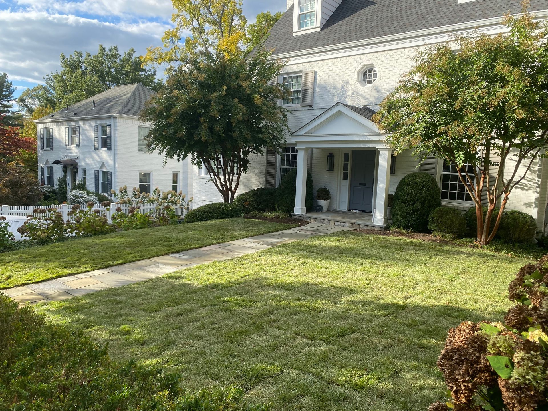 A white, two-story colonial house with a dark roof and a front porch, viewed from a sunny lawn.