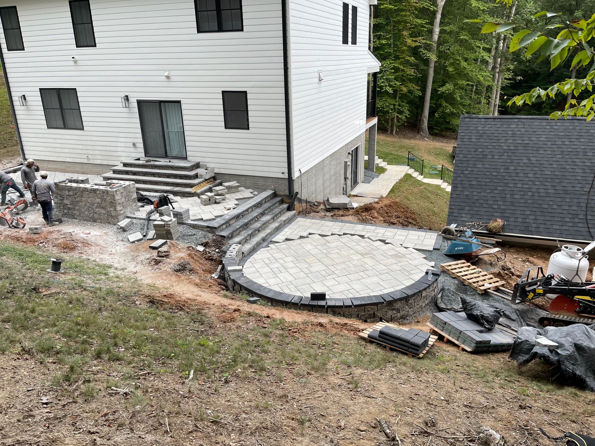 Workers build stone stairs and a circular paver patio outside the walk-out basement of a white two-story house.