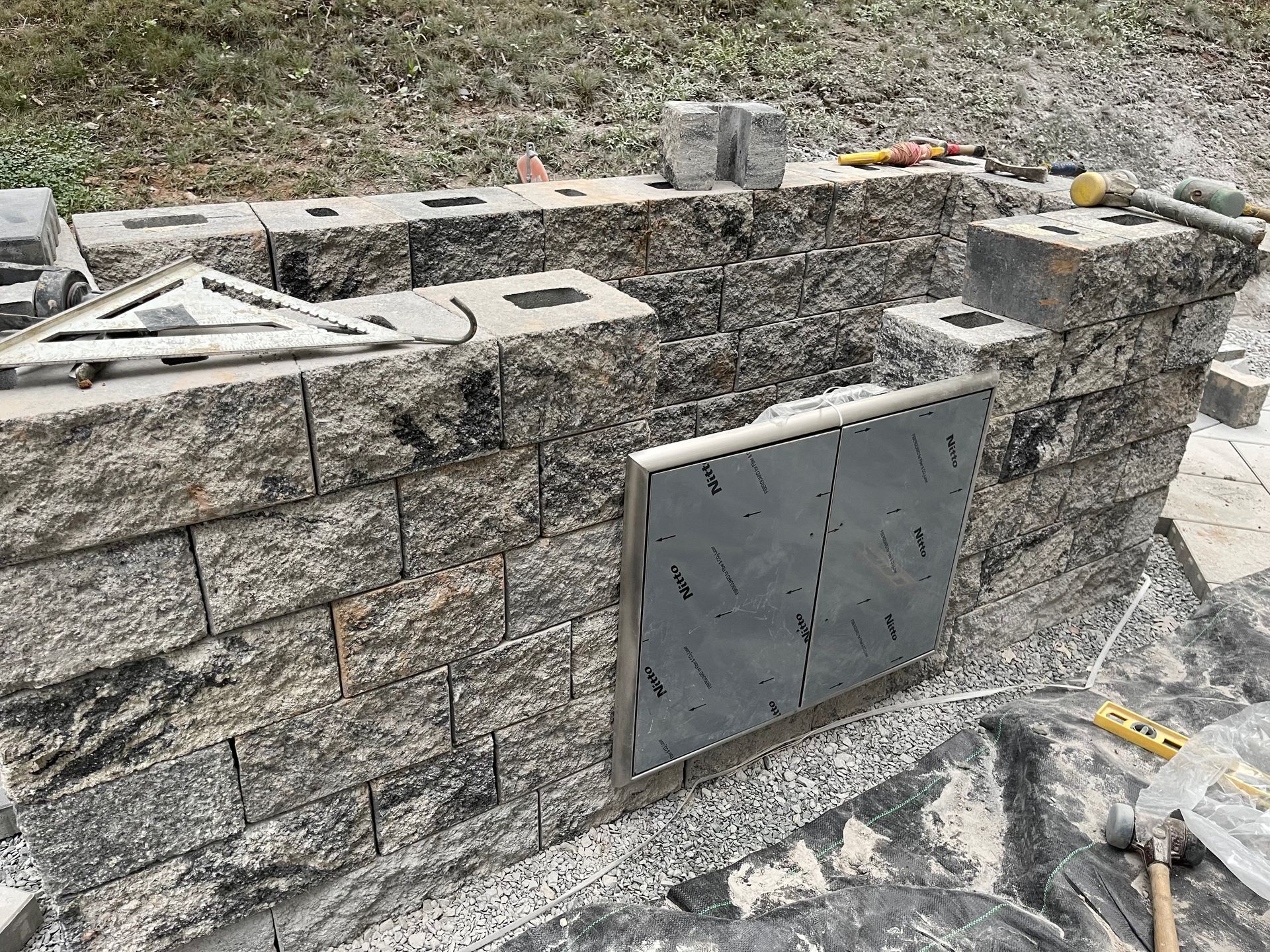 A half-built outdoor stone kitchen structure with a silver access door and construction tools on a gravel surface.