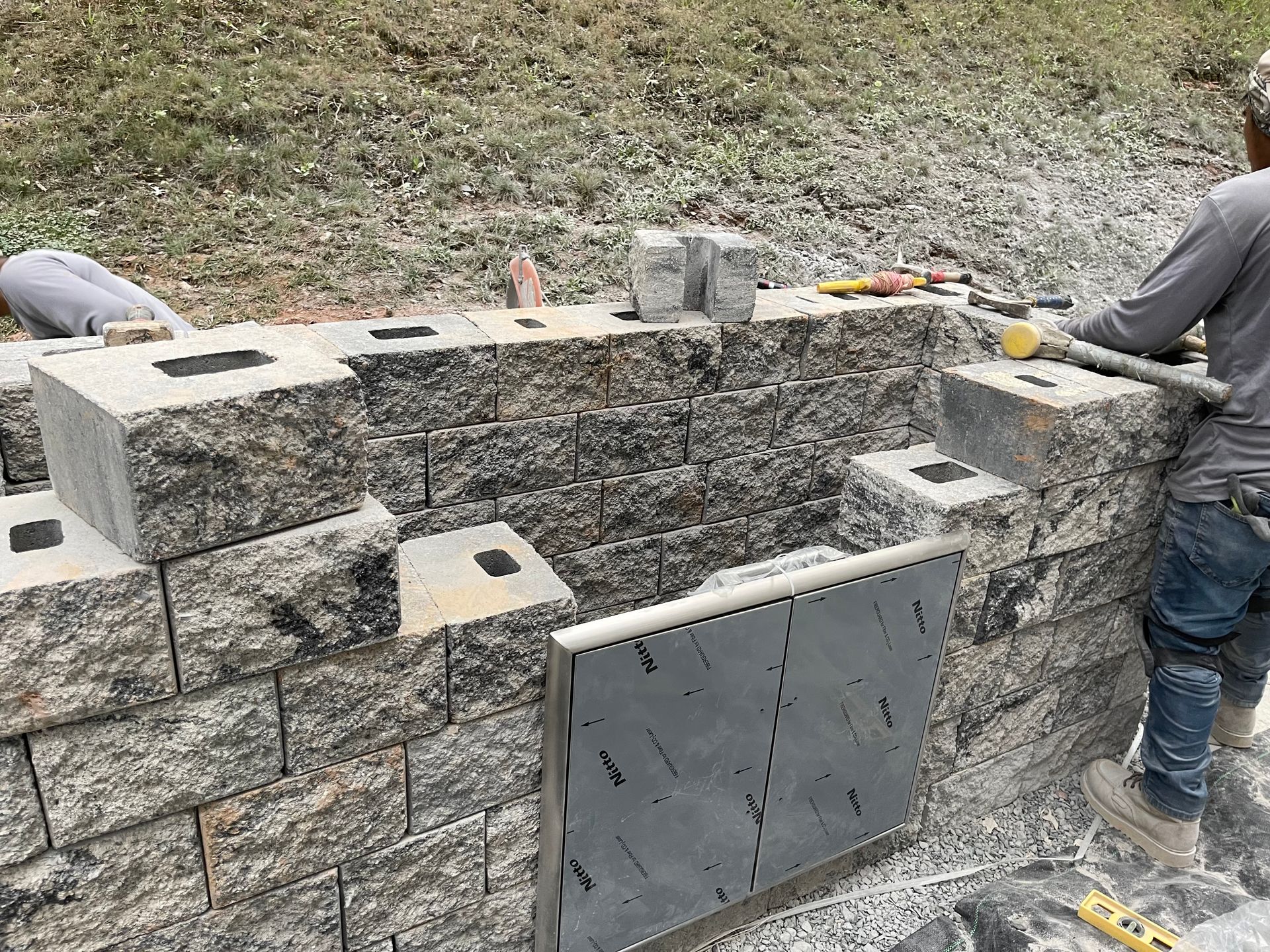 A person constructs an outdoor stone masonry wall or fireplace, using grey blocks and metal components near a grassy slope.