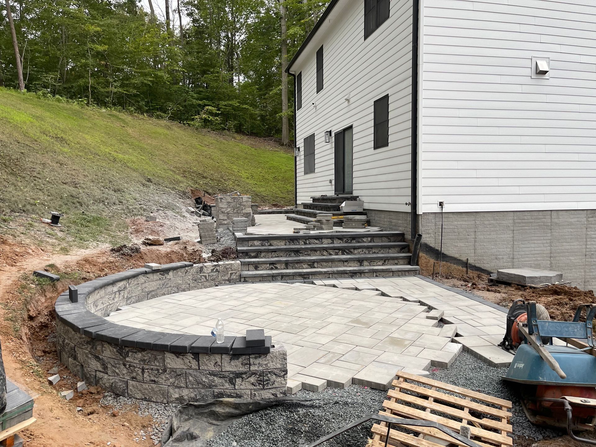 A new stone patio and stairs under construction next to a white house with a landscaped hillside in the background.