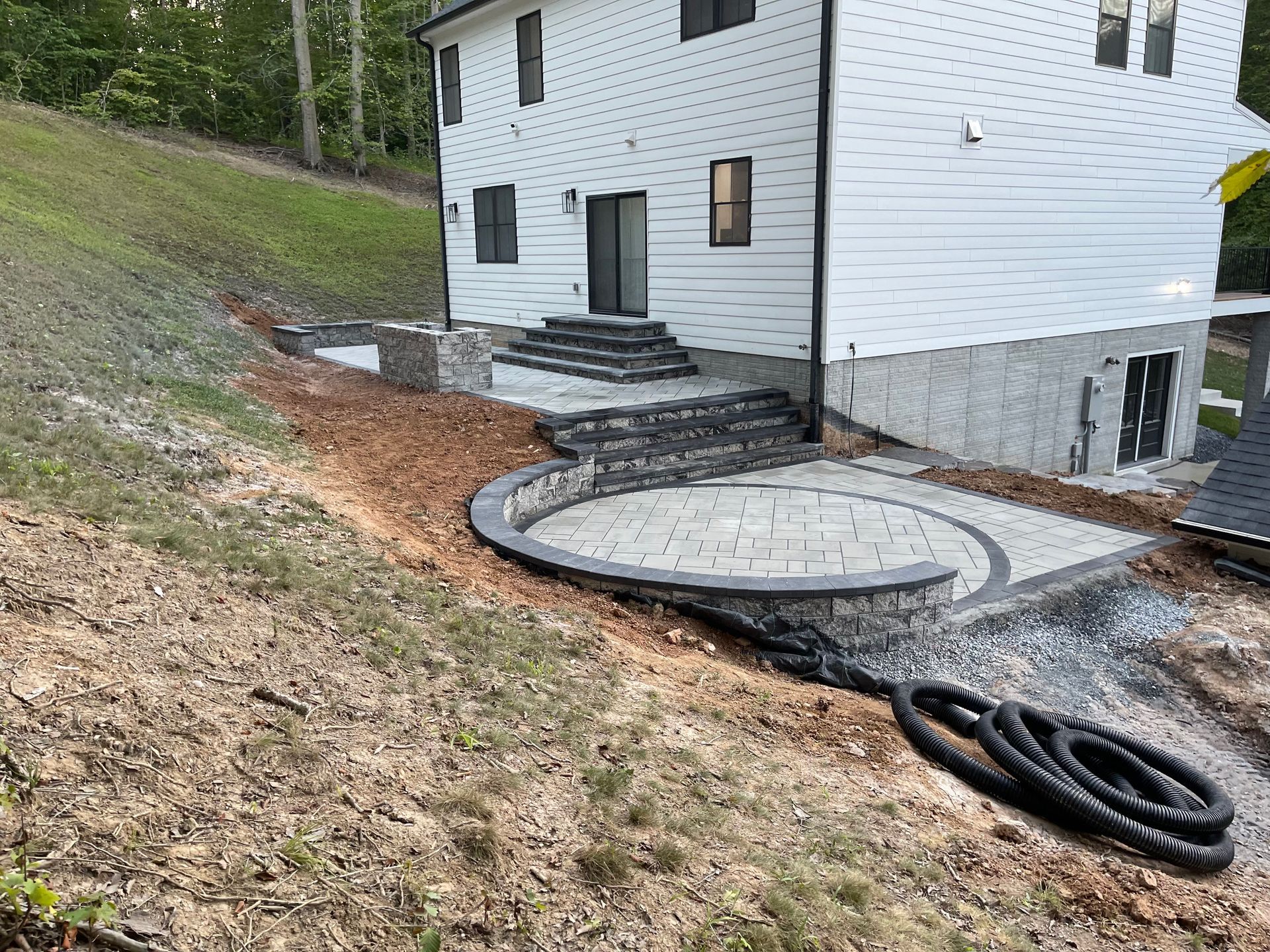 A white house with a newly installed gray stone patio and stairs leading down a sloped, grassy backyard.
