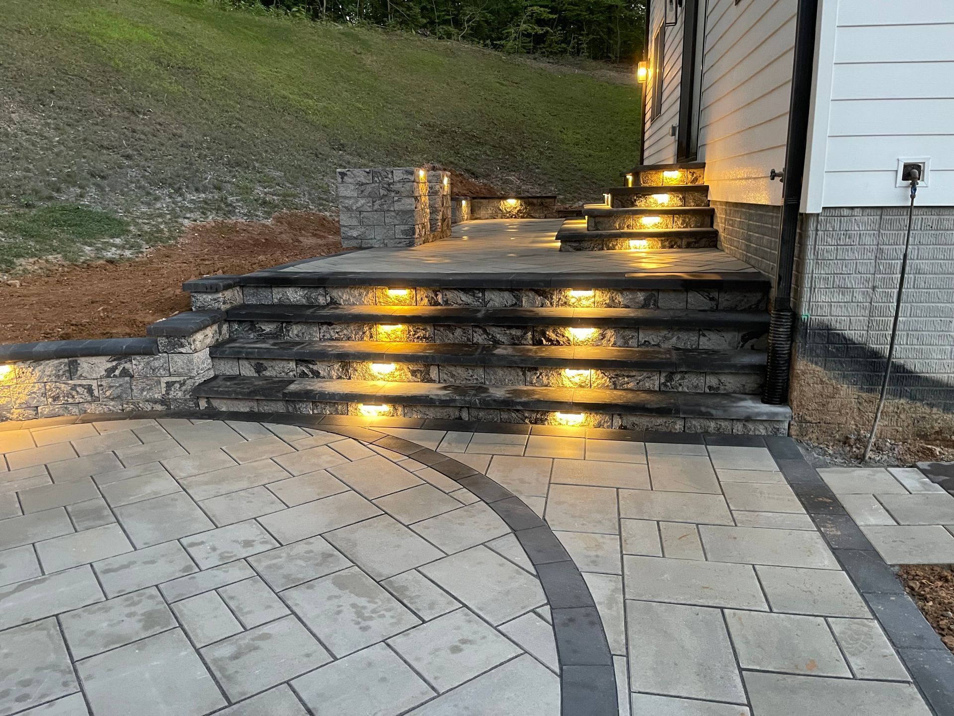 A stone patio leads to steps with integrated yellow lighting, set against a house exterior at dusk.