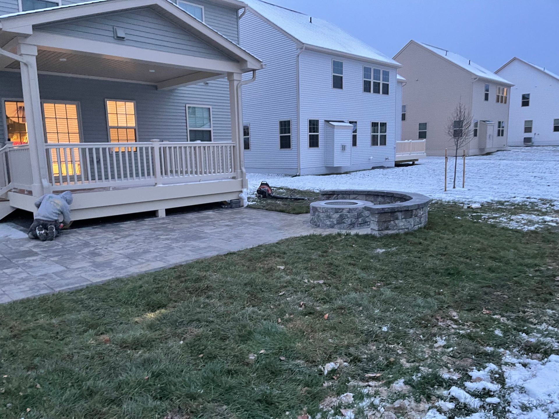 A backyard patio with a stone fire pit next to a covered deck on a light snowy day with houses in the background.