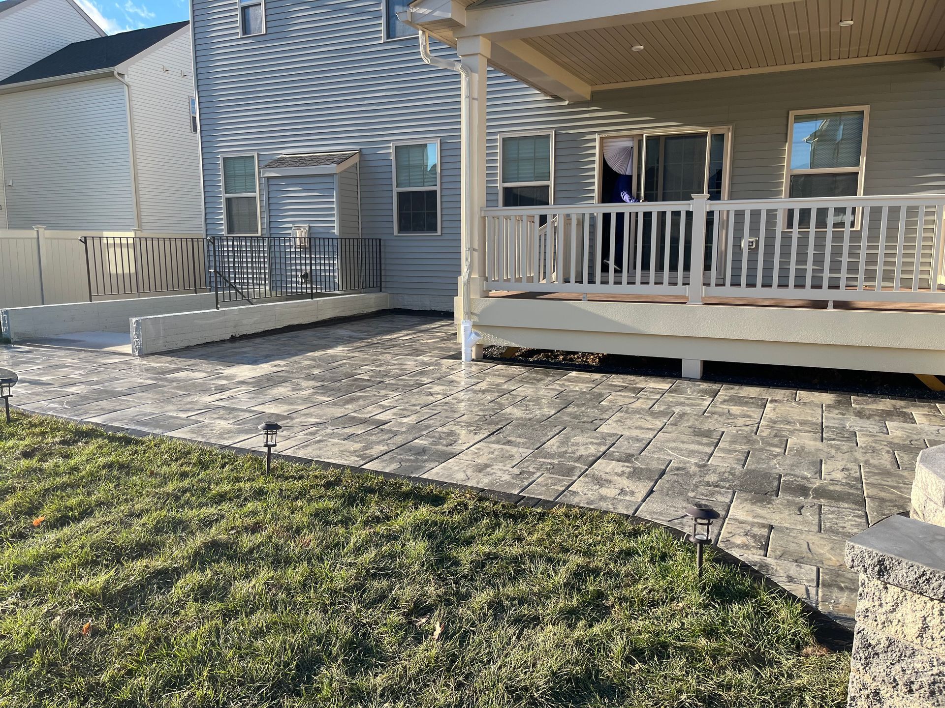 A backyard scene featuring a paved stone patio, a covered wooden deck, and a modern two-story house with gray siding.