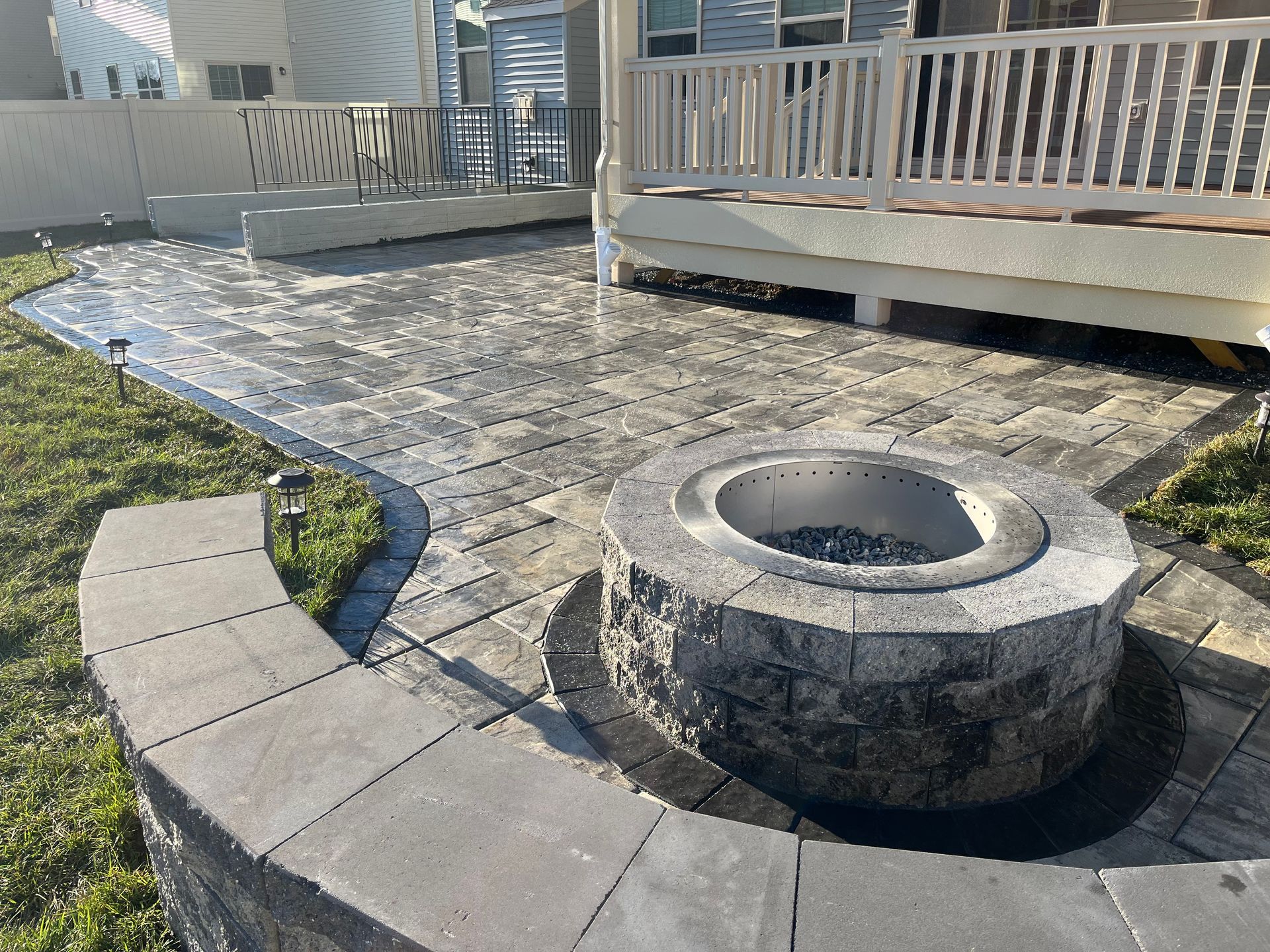 A gray paver patio with a circular stone fire pit and a curved seating wall, located next to a raised deck in a backyard.