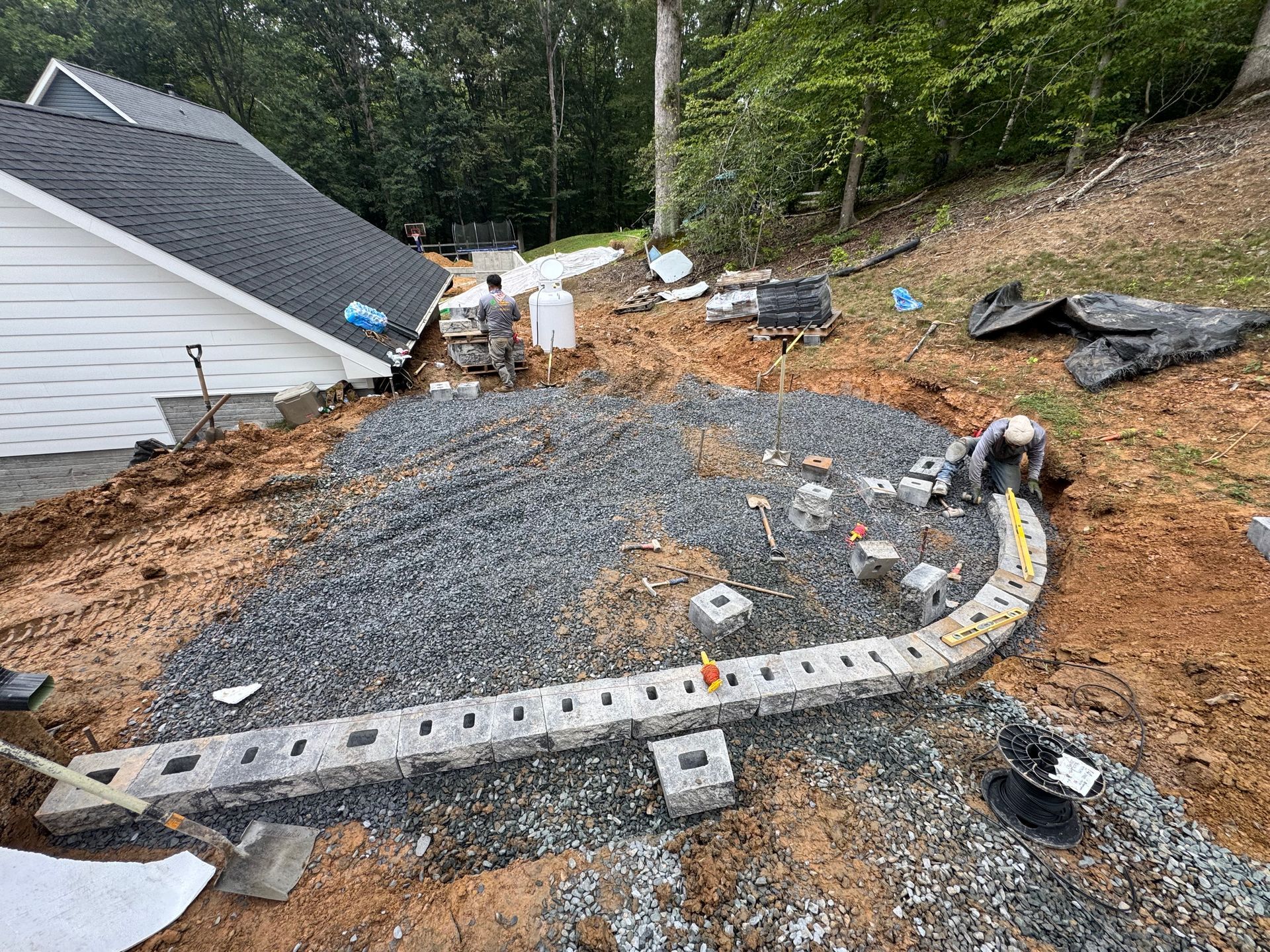 A construction worker builds a curved retaining wall using concrete blocks on a gravel-filled slope next to a house.