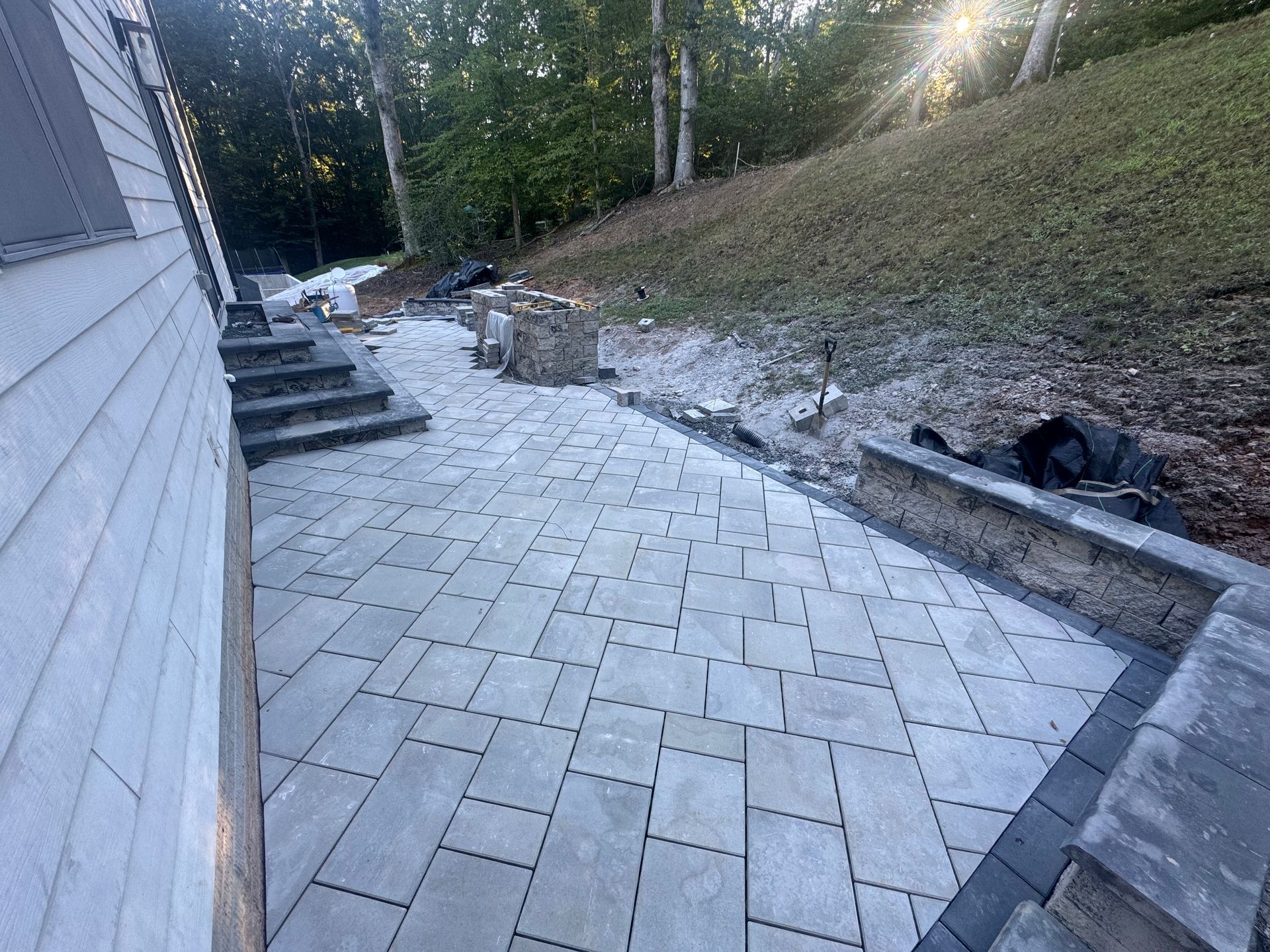 A newly installed stone paver patio next to a white-sided house with a small retaining wall and steps leading to the lawn.