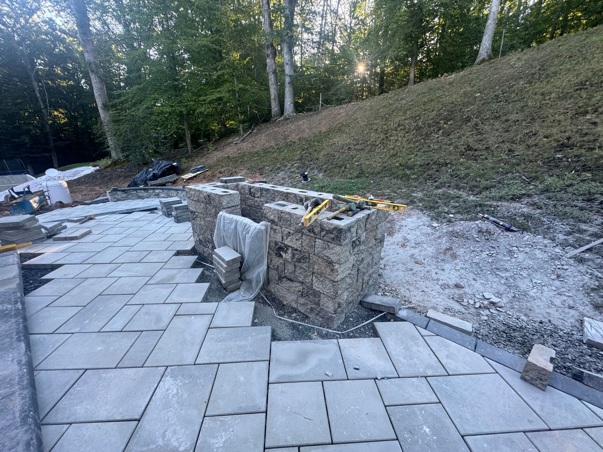 A half-built stone pillar and outdoor patio under construction with rectangular grey stone pavers on a wooded hillside.