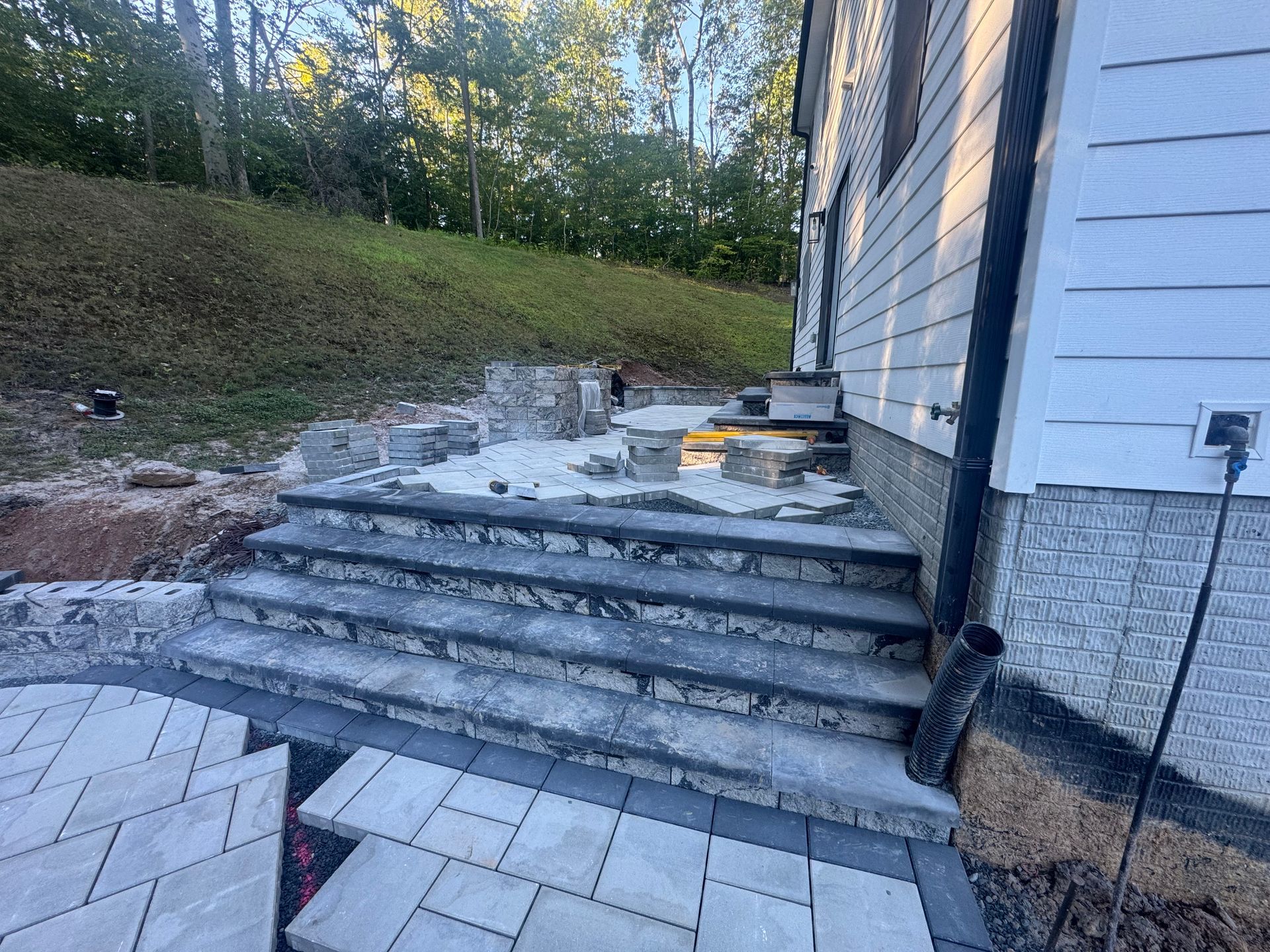 Newly built stone patio stairs leading to a back door and a half-finished stone patio area with trees in the background.