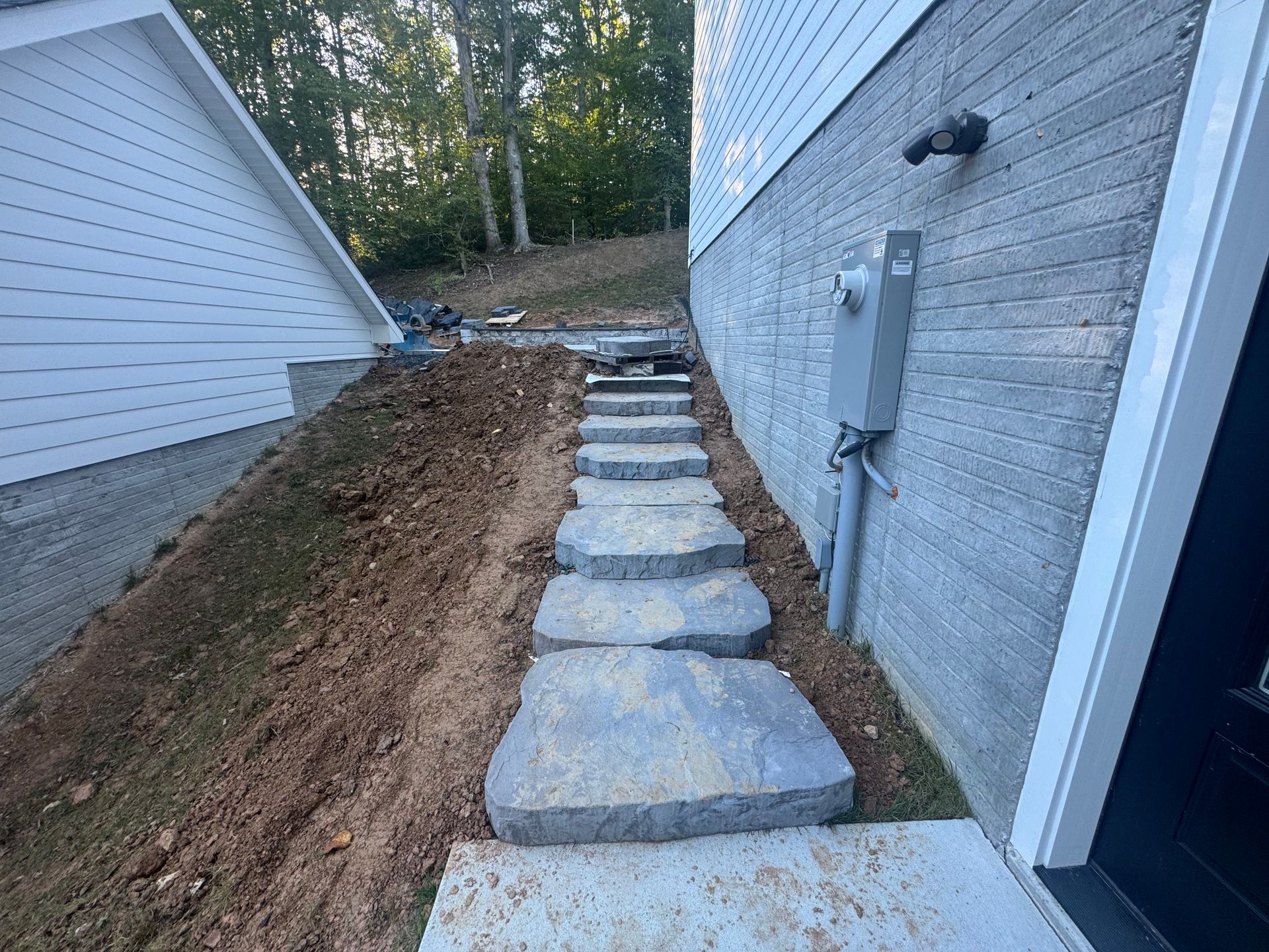 A stone stairway between a house wall and an earthen slope leads upward toward a wooded area.