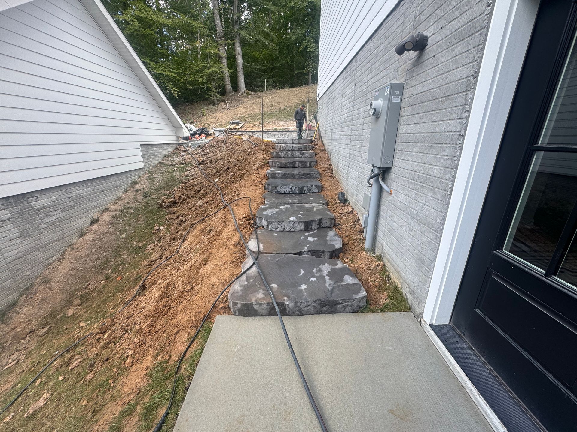 Stone steps ascend a grassy slope between two house walls, leading from a concrete walkway toward a wooded area.
