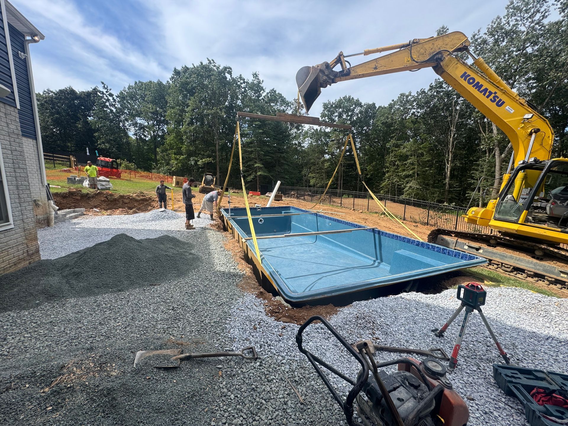 A yellow excavator lowers a pre-molded blue swimming pool into a backyard excavation site filled with gravel.