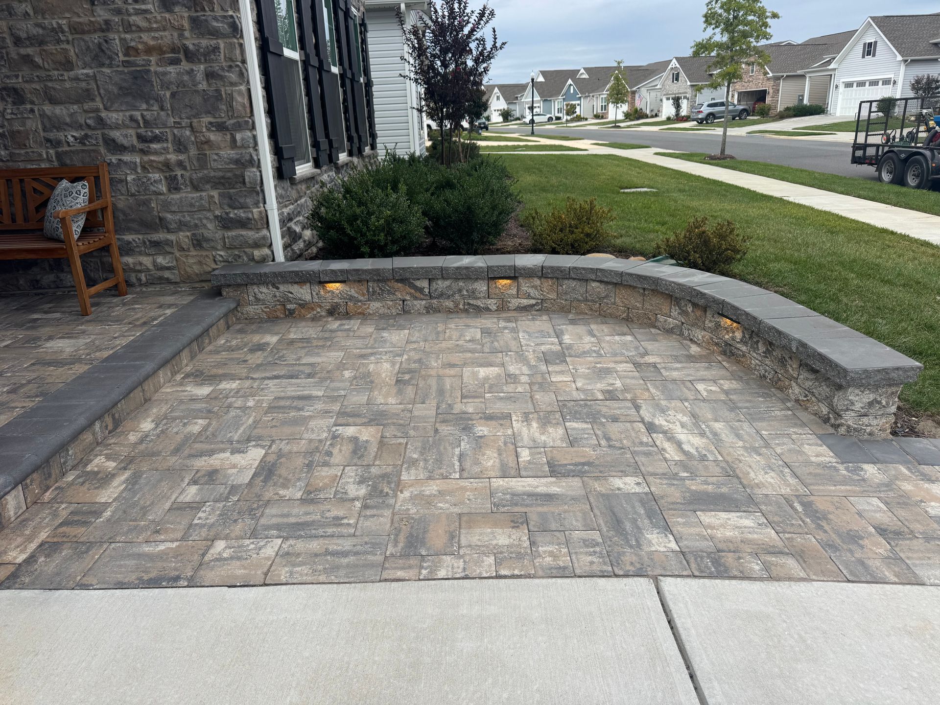 A stone paver patio with a curved seating wall featuring built-in lighting in a suburban residential front yard.