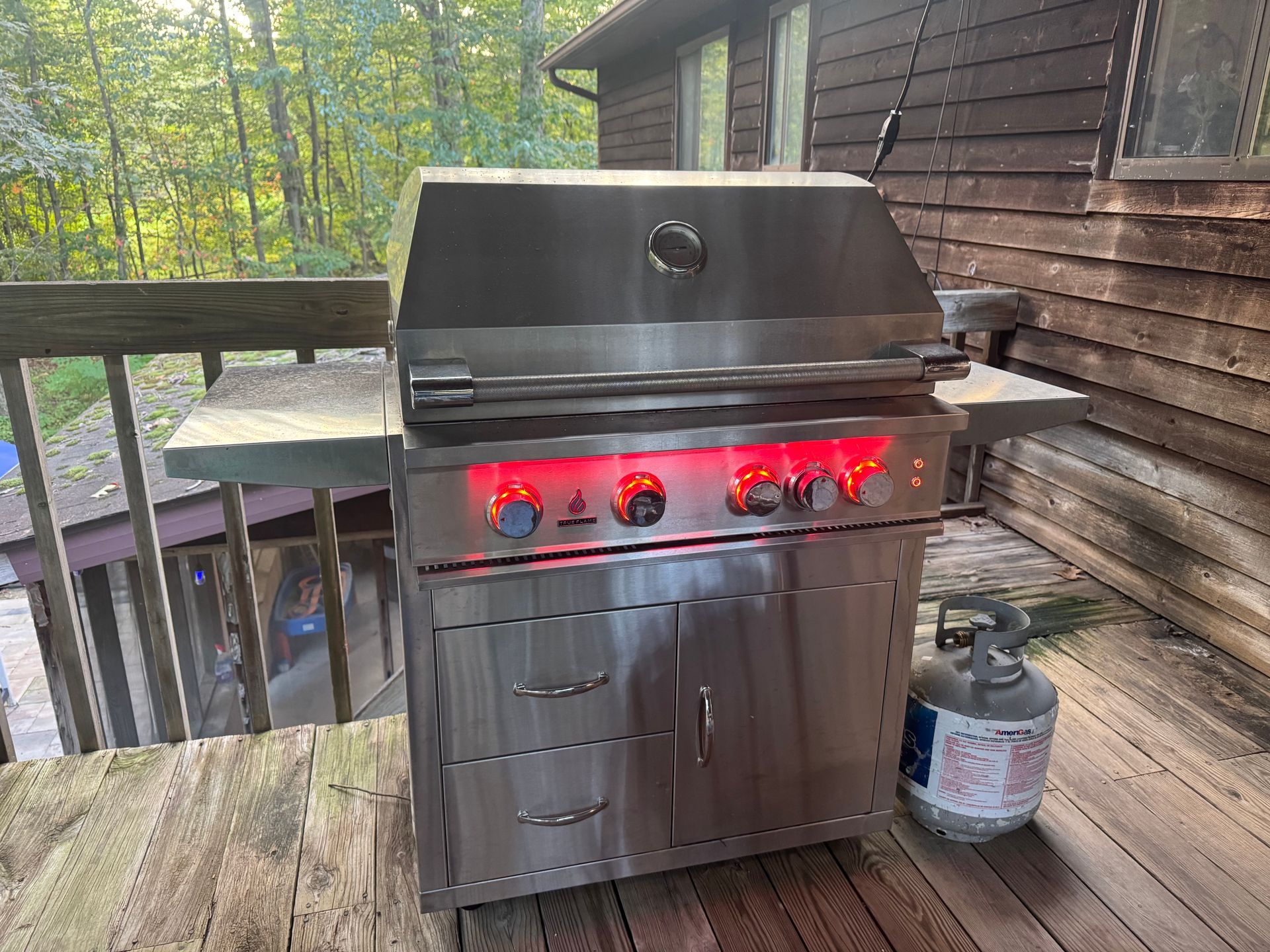 A stainless steel gas grill with illuminated red knobs sits on a wooden deck next to a propane tank.