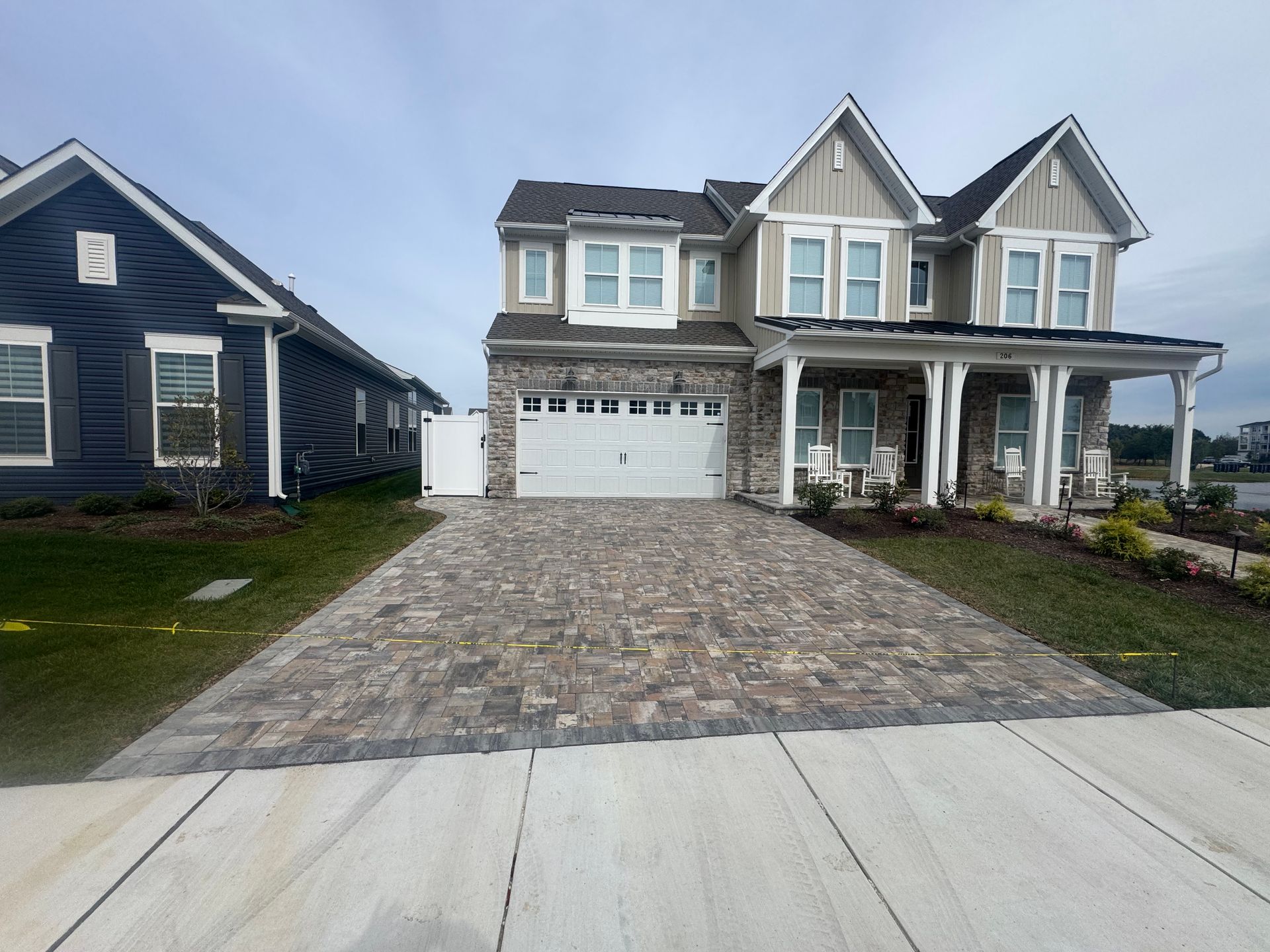 A two-story tan home with stone accents and a patterned paver driveway sits beside a dark blue house under a cloudy sky.