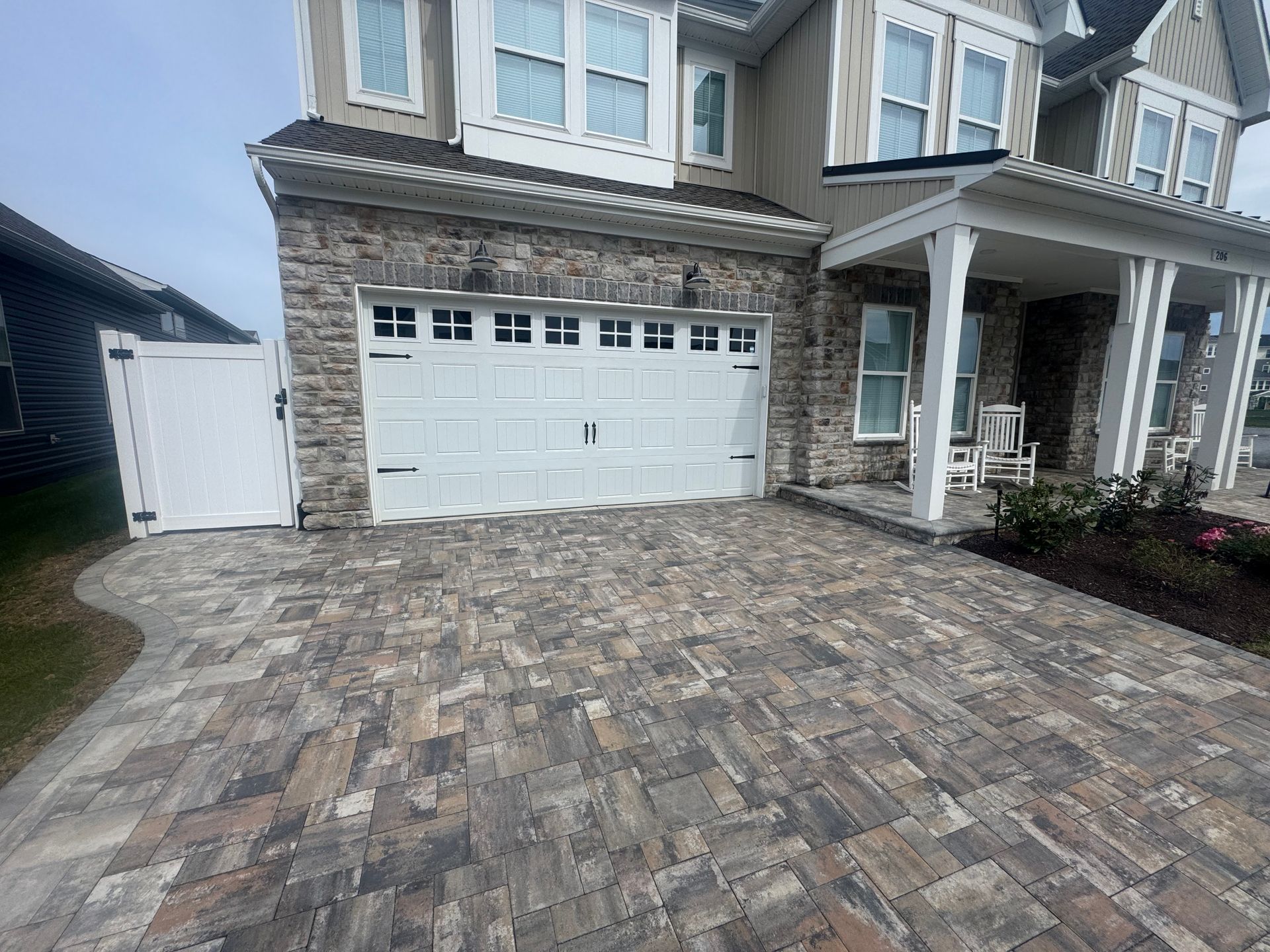 A paved driveway leads to a stone-front house with a white garage door, a front porch, and a white side gate.
