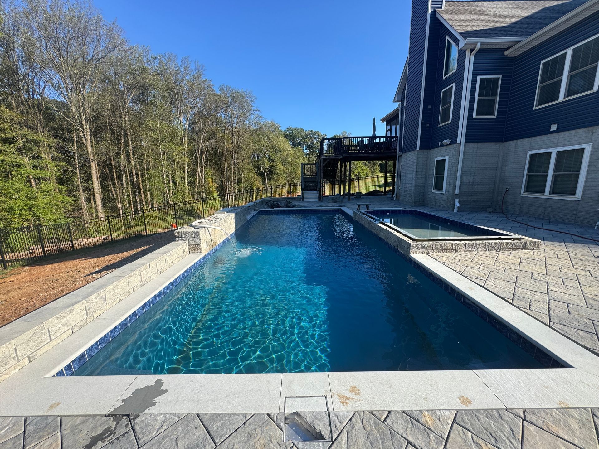 A rectangular blue swimming pool with an attached spa next to a dark blue house and a stone patio on a sunny day.
