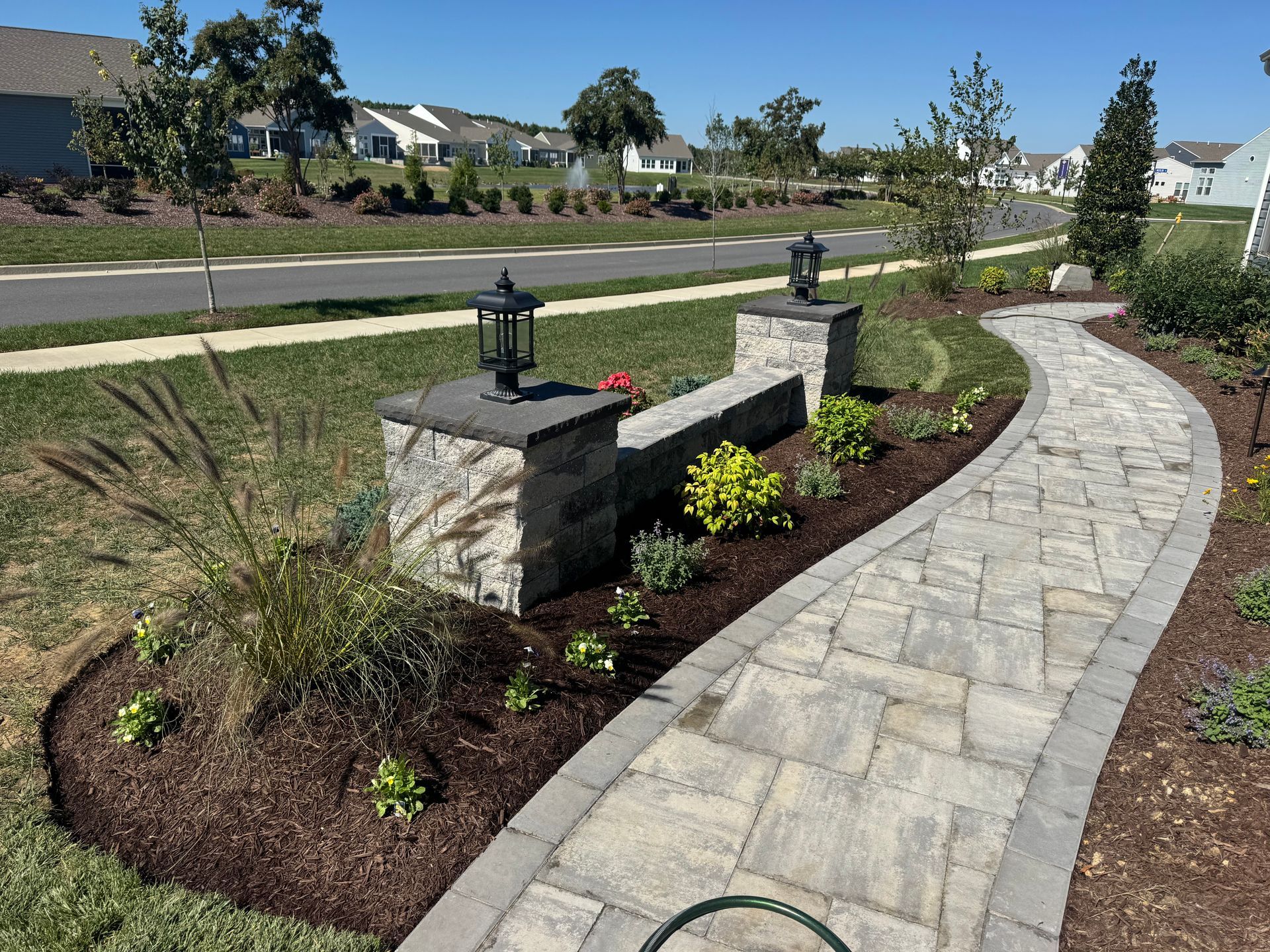 A winding stone path leads past a low stone retaining wall topped with two black light fixtures in a suburban landscape.