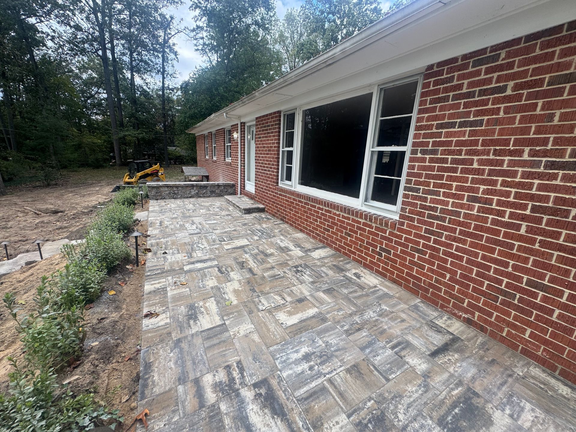 A newly installed gray paver patio extends along the side of a red brick house, next to a landscaped garden bed.