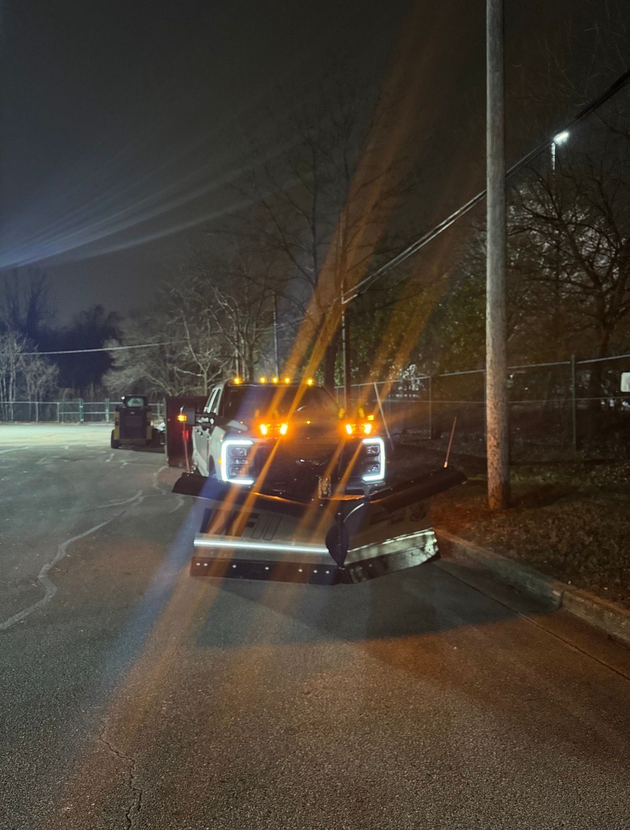 A truck equipped with a snow plow parked at night next to a utility pole in a paved lot.