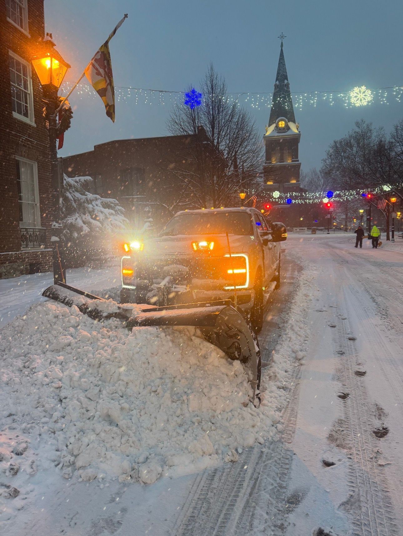 A truck with a plow clearing deep snow from a road in a town square at dusk, with a church tower in the background.