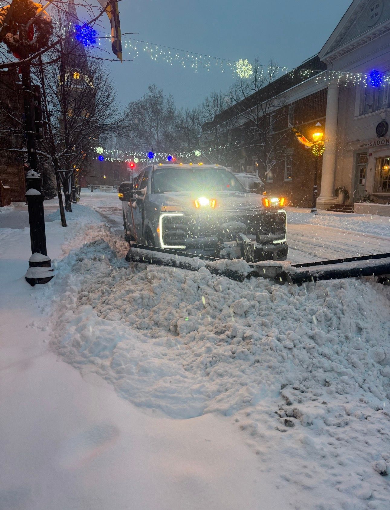 A pickup truck with its headlights on sits in a snowy street near a downed utility pole and string lights at dusk.