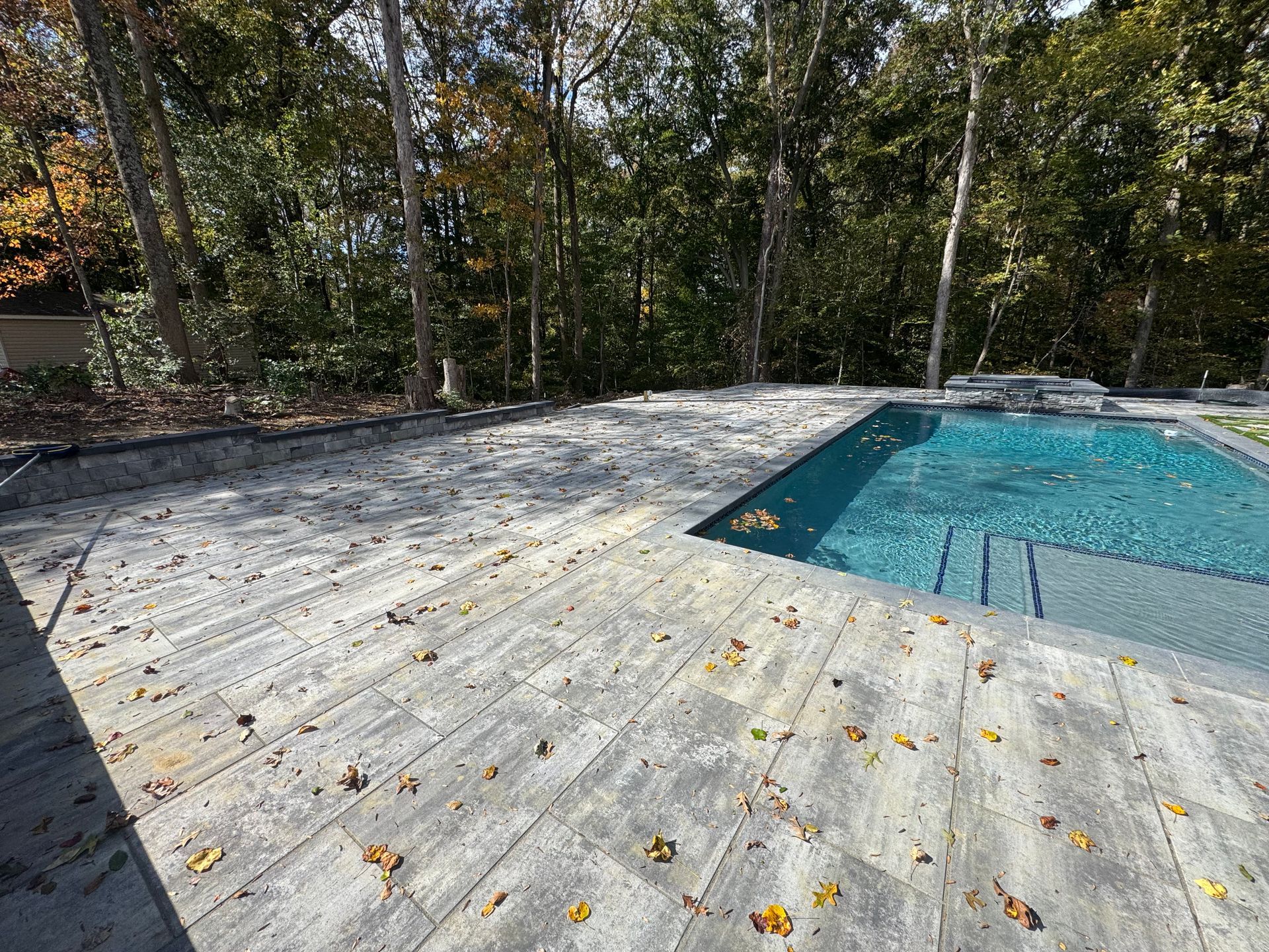 A stone patio with scattered autumn leaves beside a rectangular blue swimming pool, set against a backdrop of trees.