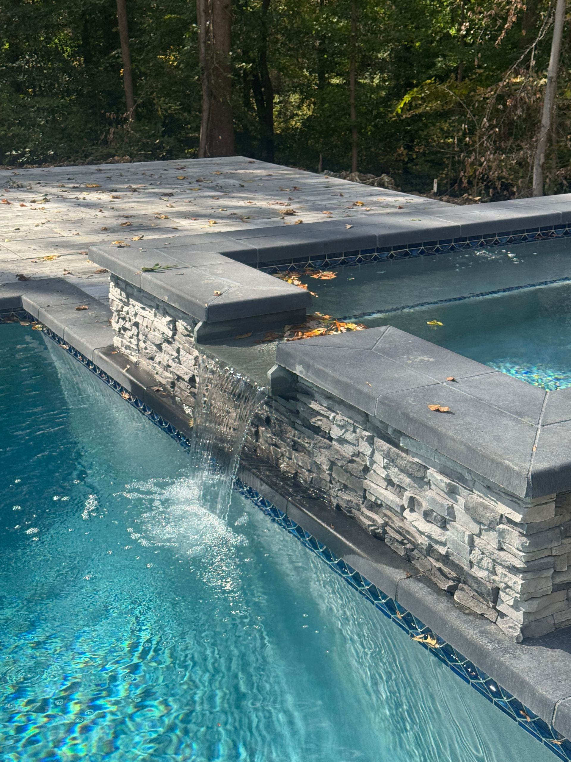 A tiered swimming pool with a stone-accented spillway, dark gray coping, and clear blue water set in a wooded area.