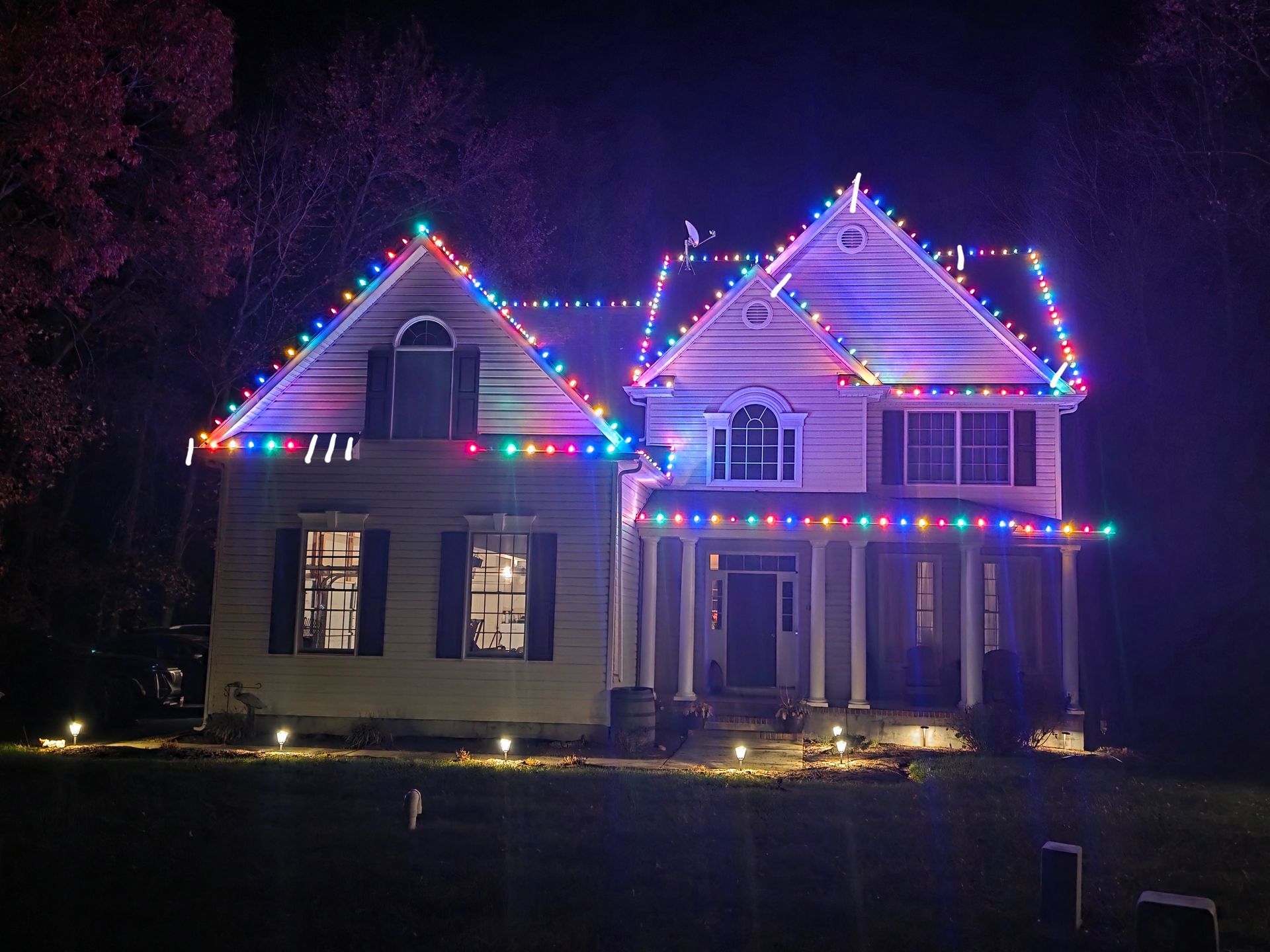 A two-story house at night outlined with vibrant, multicolored holiday string lights against a dark, treed backdrop.