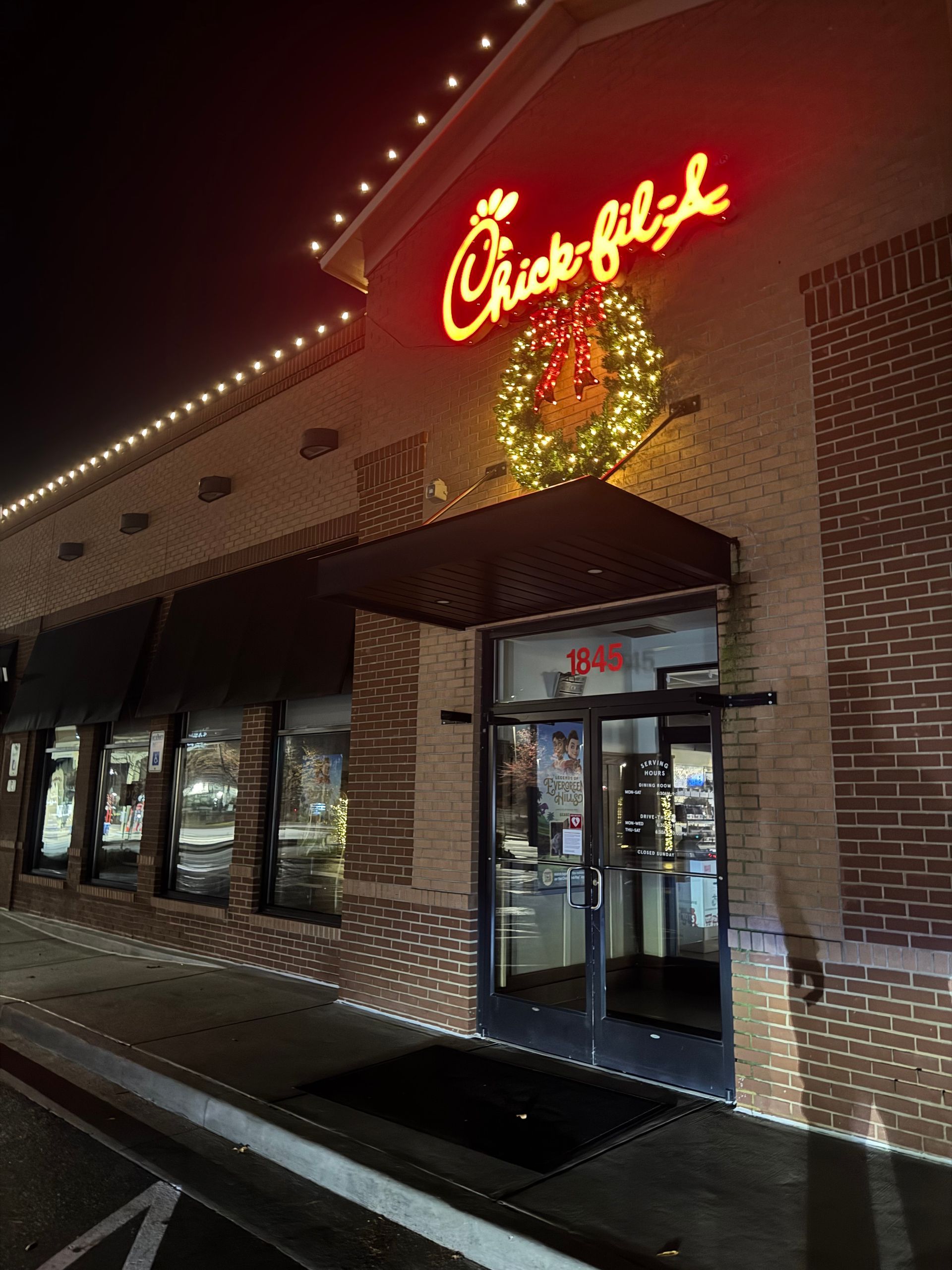 Exterior of a brick Chick-fil-A restaurant at night, decorated with a glowing wreath and white string lights.
