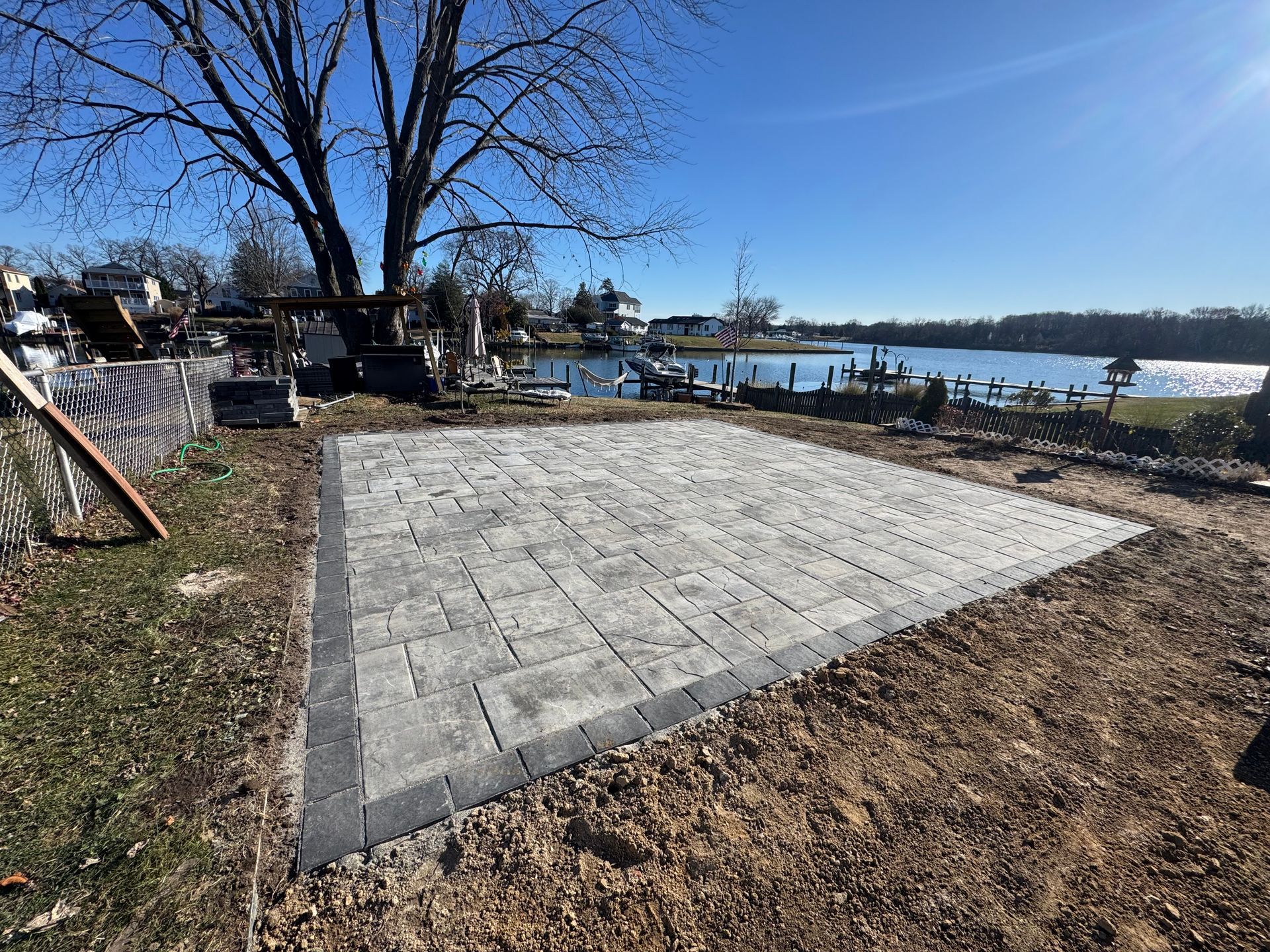 A square patio made of grey and black pavers sits in a yard overlooking a lake under a clear blue sky.