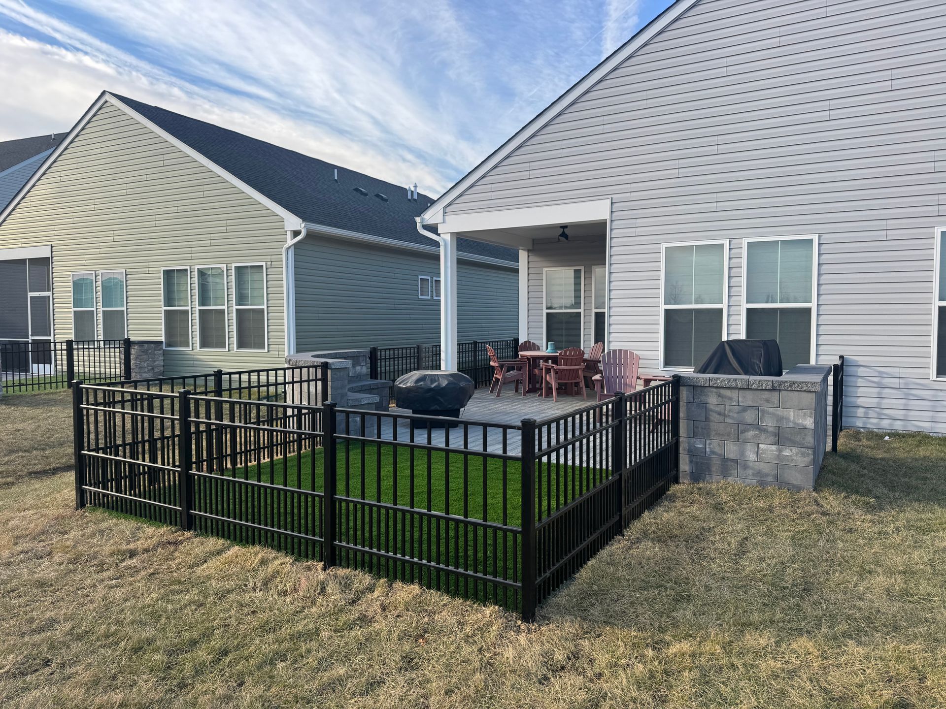 A backyard patio with a black metal fence enclosing a small artificial grass area, adjacent to a house with gray siding.