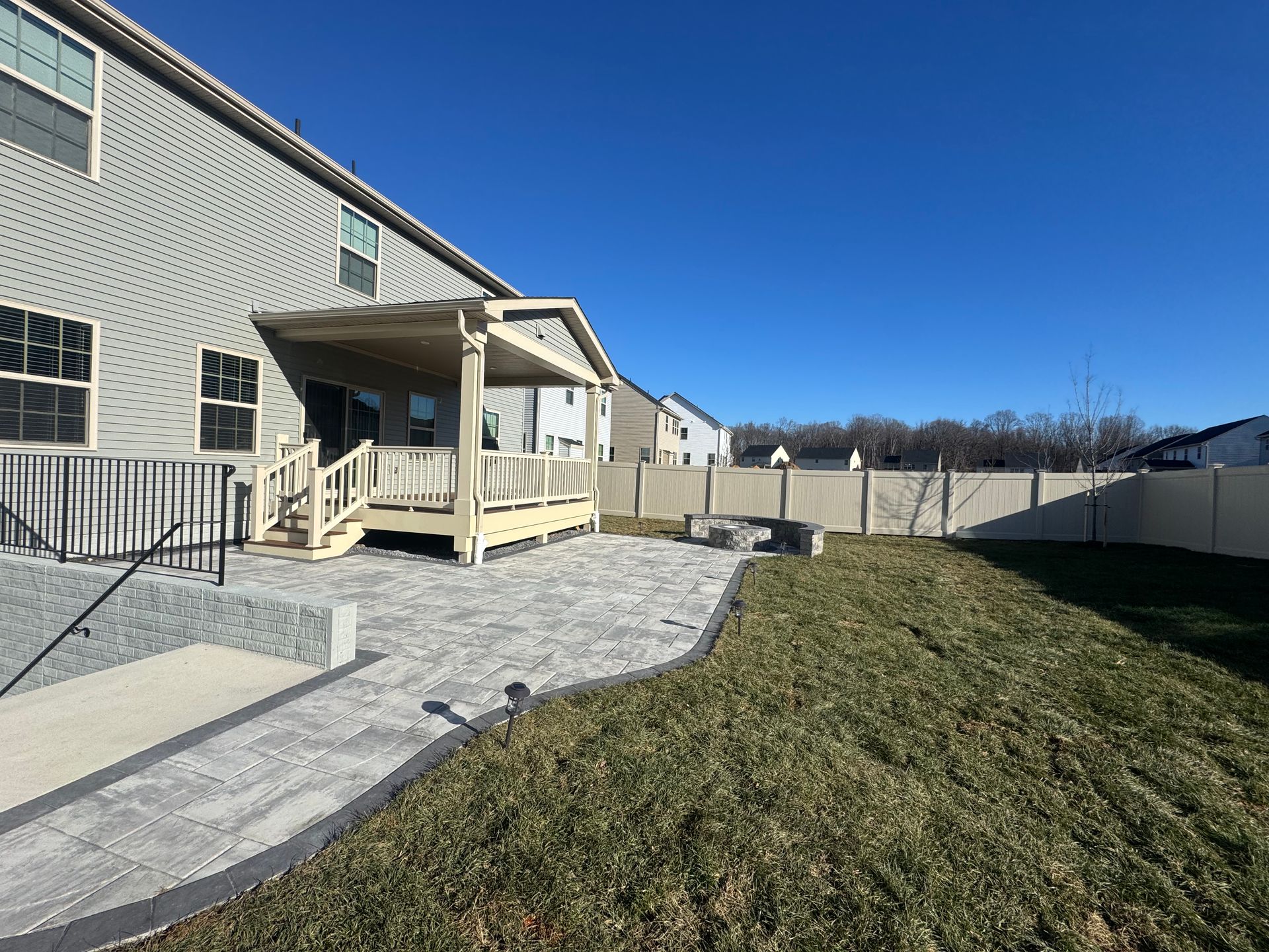 A backyard featuring a beige house with a covered patio, stone paver patio area, and a grassy lawn enclosed by a fence.