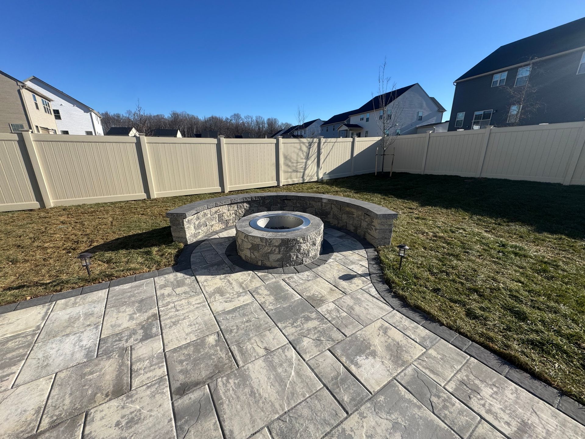 A stone fire pit with a curved seating wall on a paved patio in a suburban backyard enclosed by a beige vinyl fence.