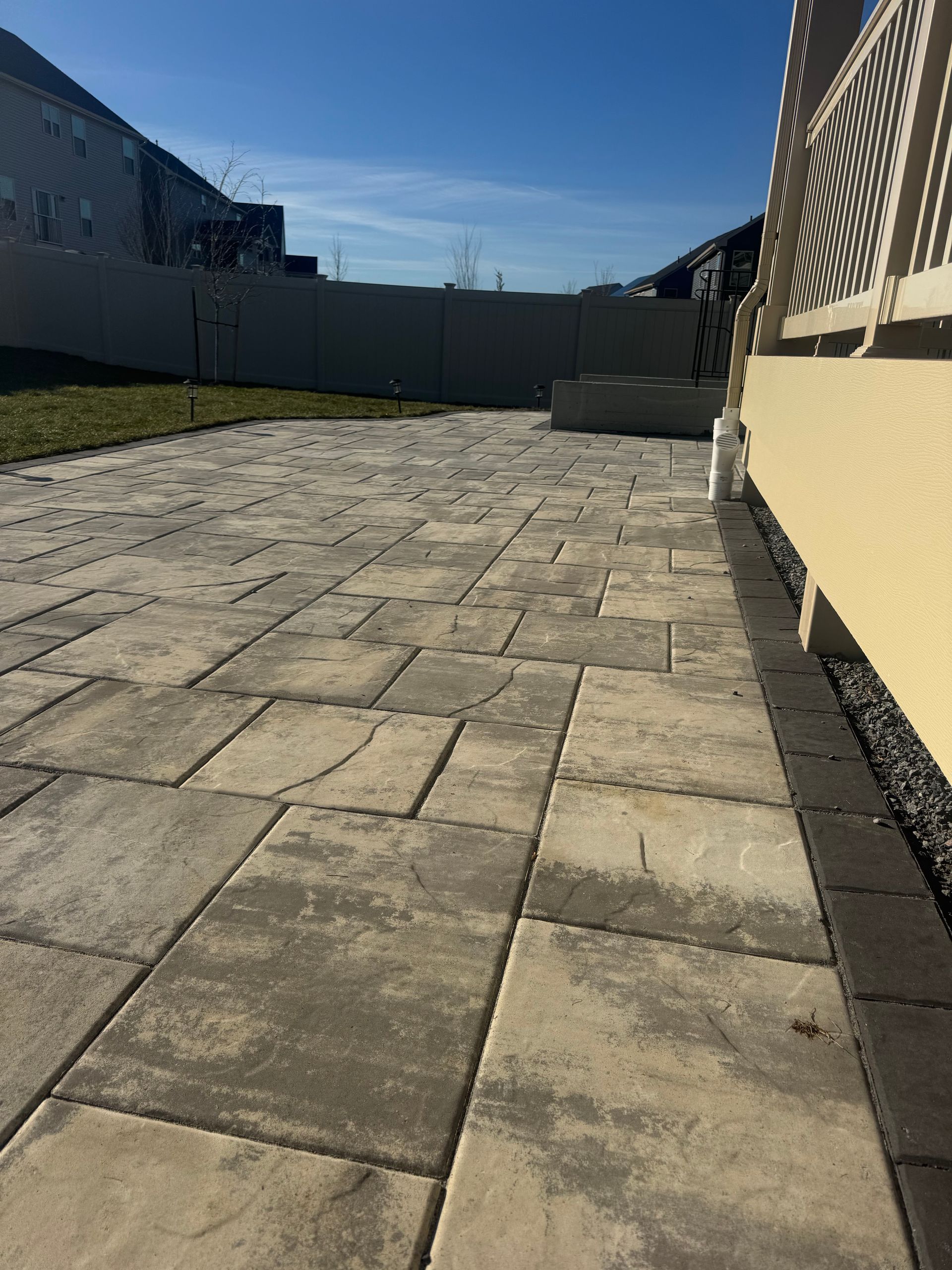 A light-colored stone patio with a dark border runs alongside a yellow building exterior under a clear blue sky.