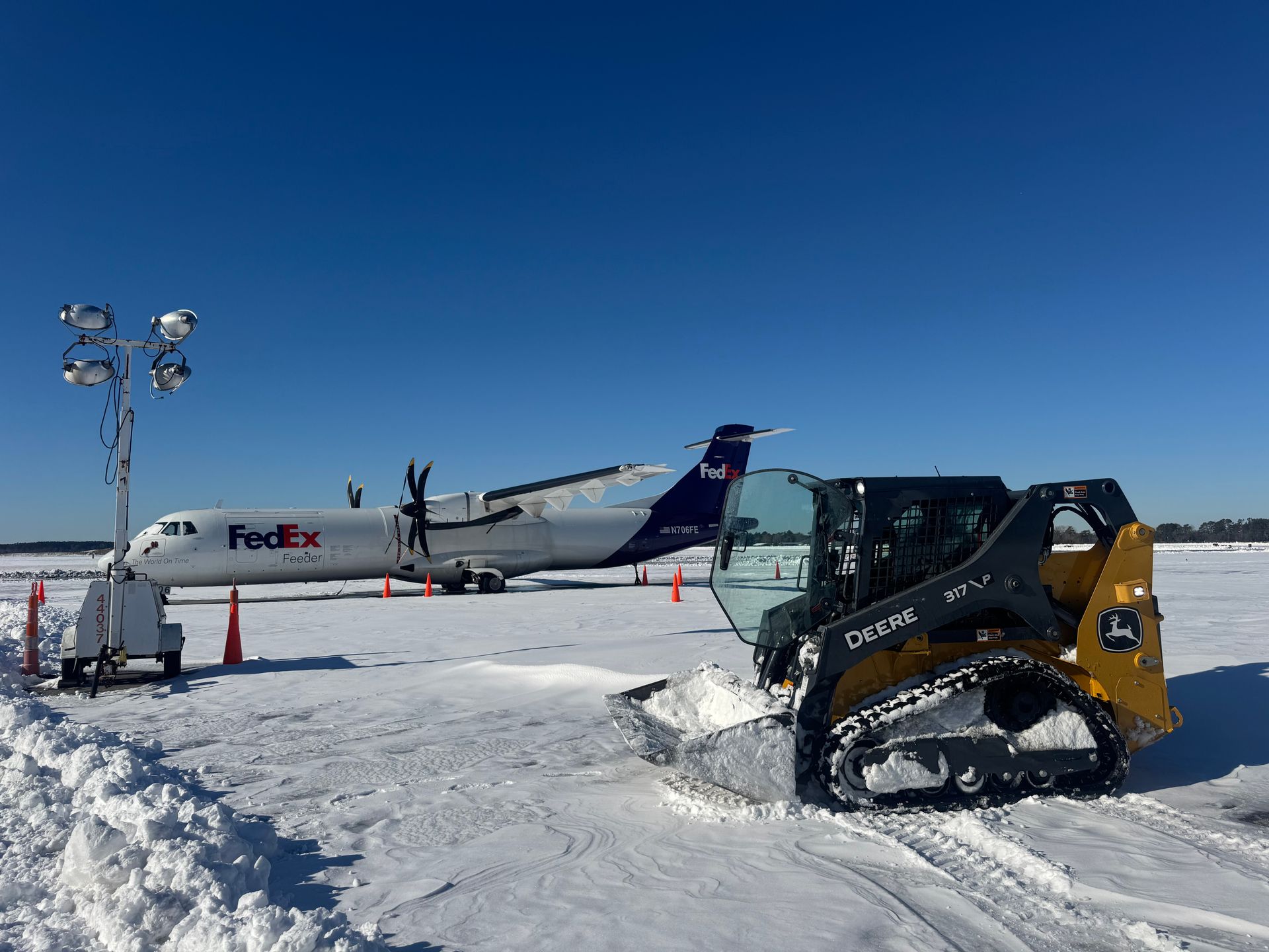 A yellow John Deere skid-steer clears snow on an airfield with a parked FedEx propeller plane and light tower in view.