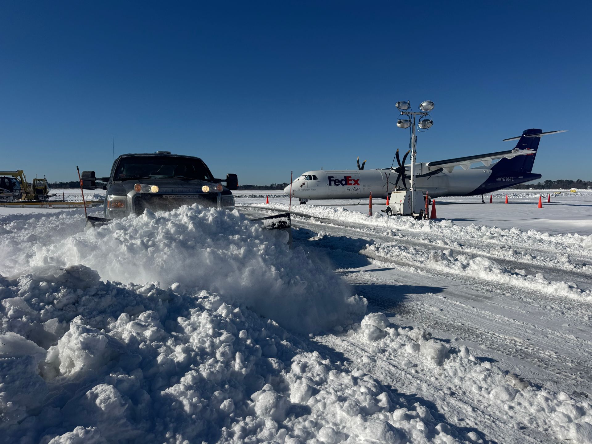 A snowplow clears a snow-covered airport tarmac near a parked FedEx cargo airplane under a bright blue sky.