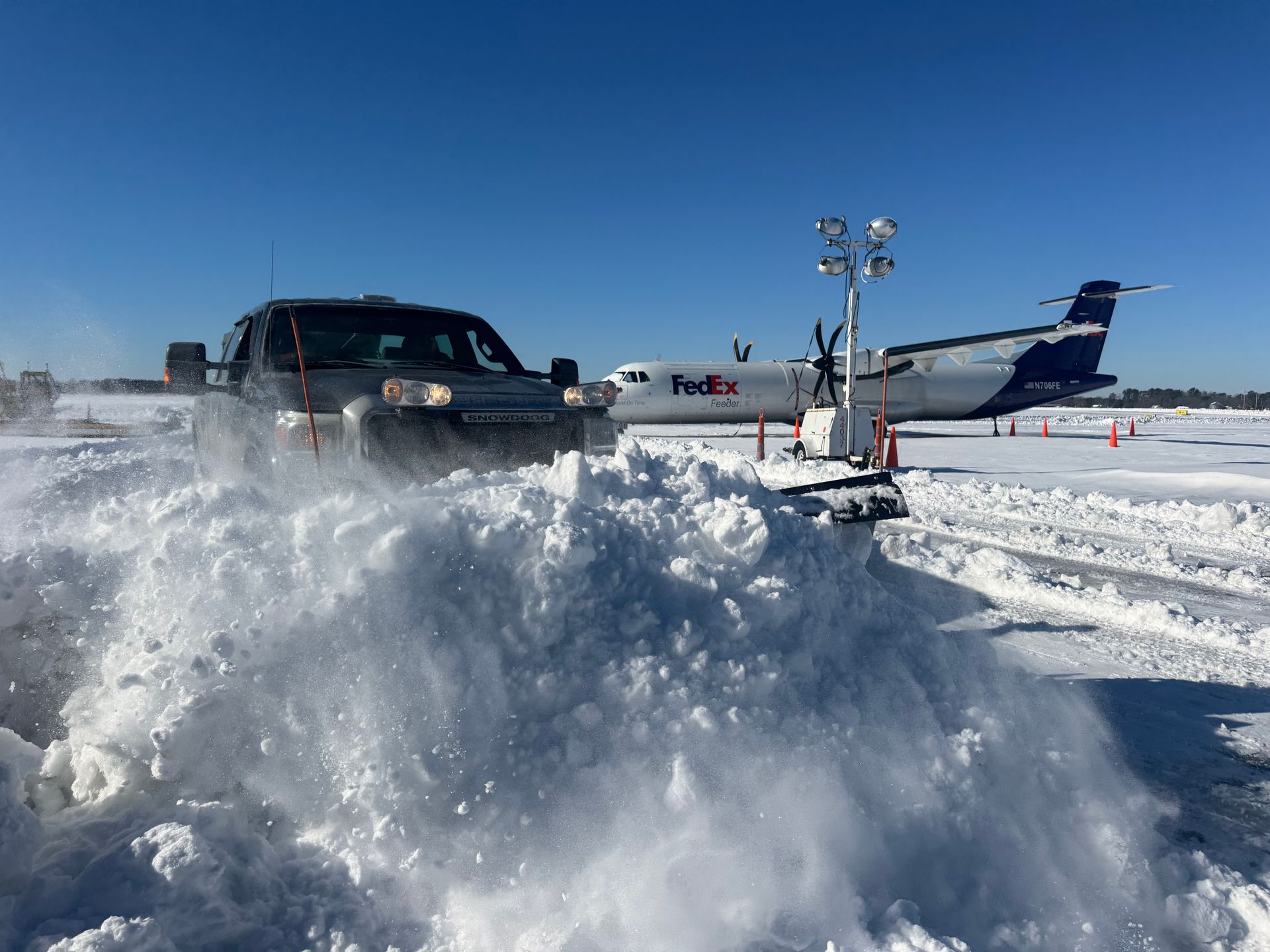 A dark pickup truck plows a large mound of snow on an airport tarmac with a FedEx airplane parked in the background.