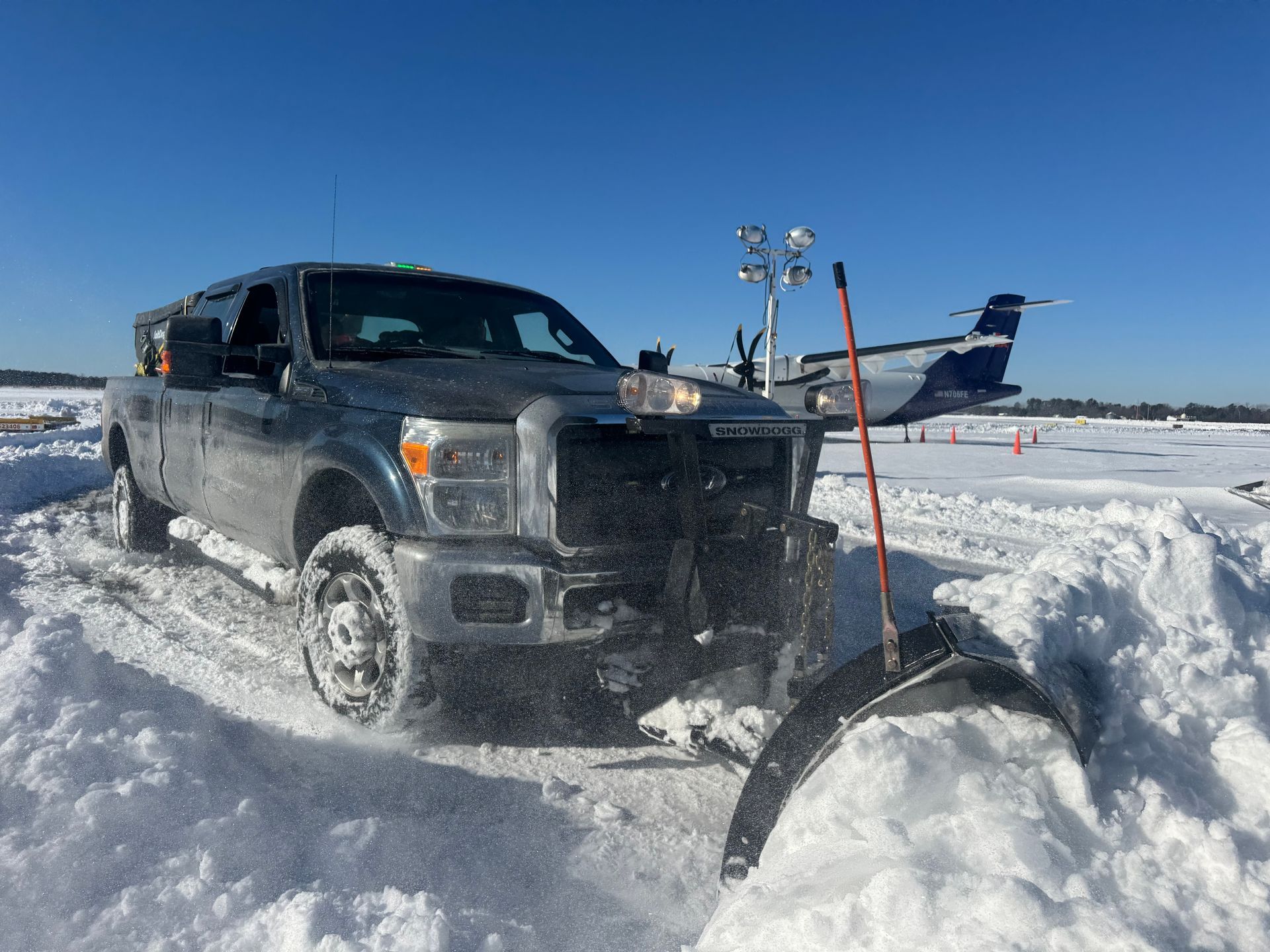 A dark-colored pickup truck with a snowplow blade clears snow on a bright, sunny day with an airplane in the background.