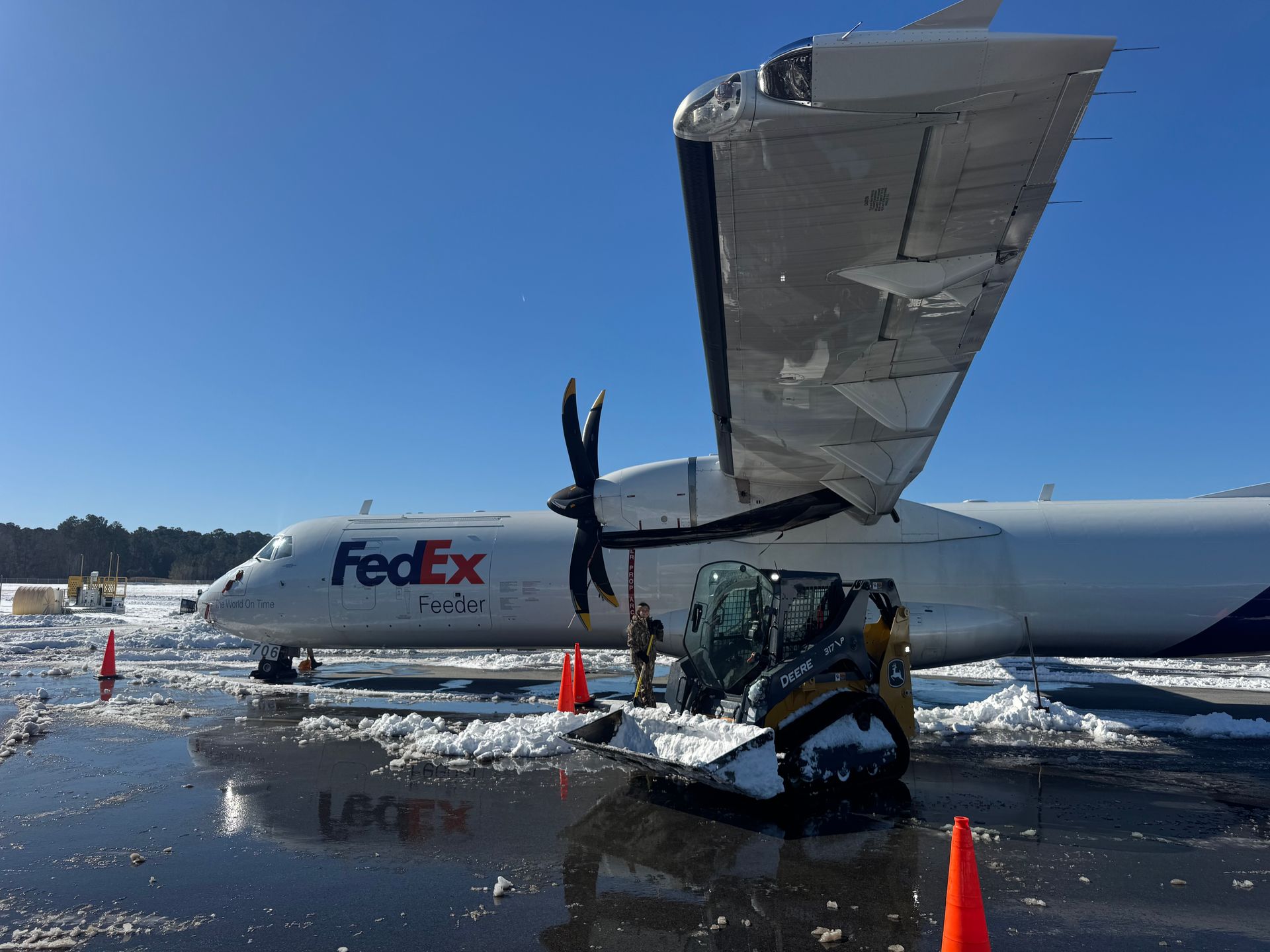 A yellow skid-steer loader clears snow near a FedEx airplane parked on a wet, icy tarmac under a clear blue sky.
